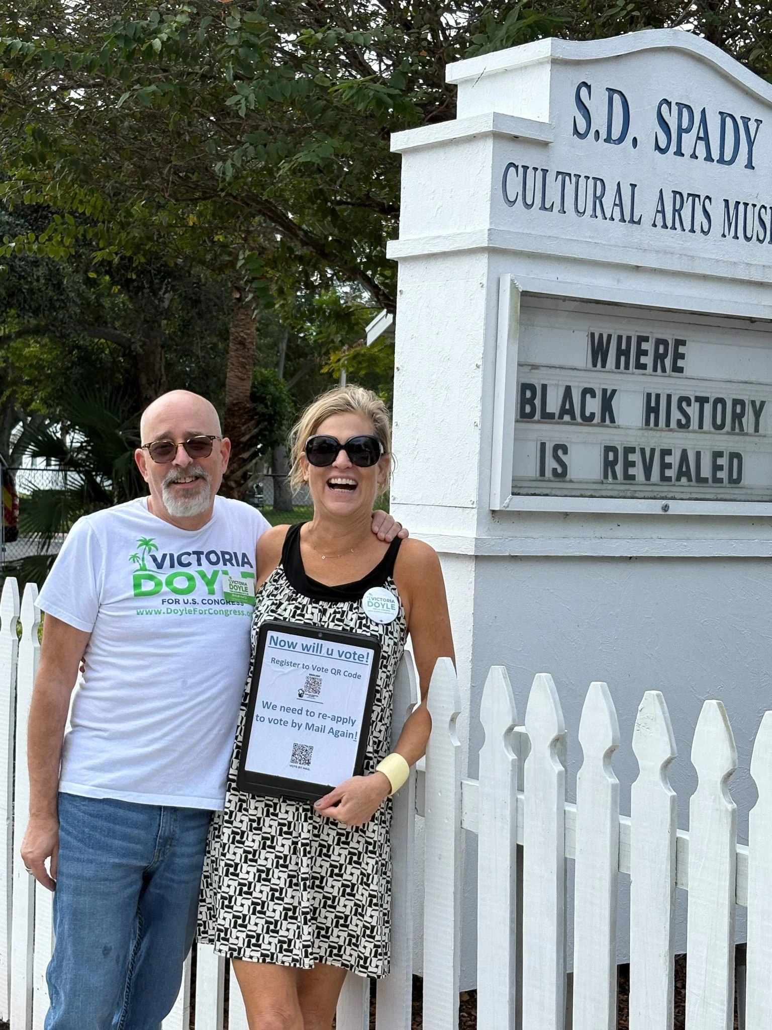 Two smiling individuals, a man and a woman, standing outside the S.D. Spady Cultural Arts Museum, holding a sign about voting, in front of a white picket fence and a sign that reads 'Where Black History is Revealed.'