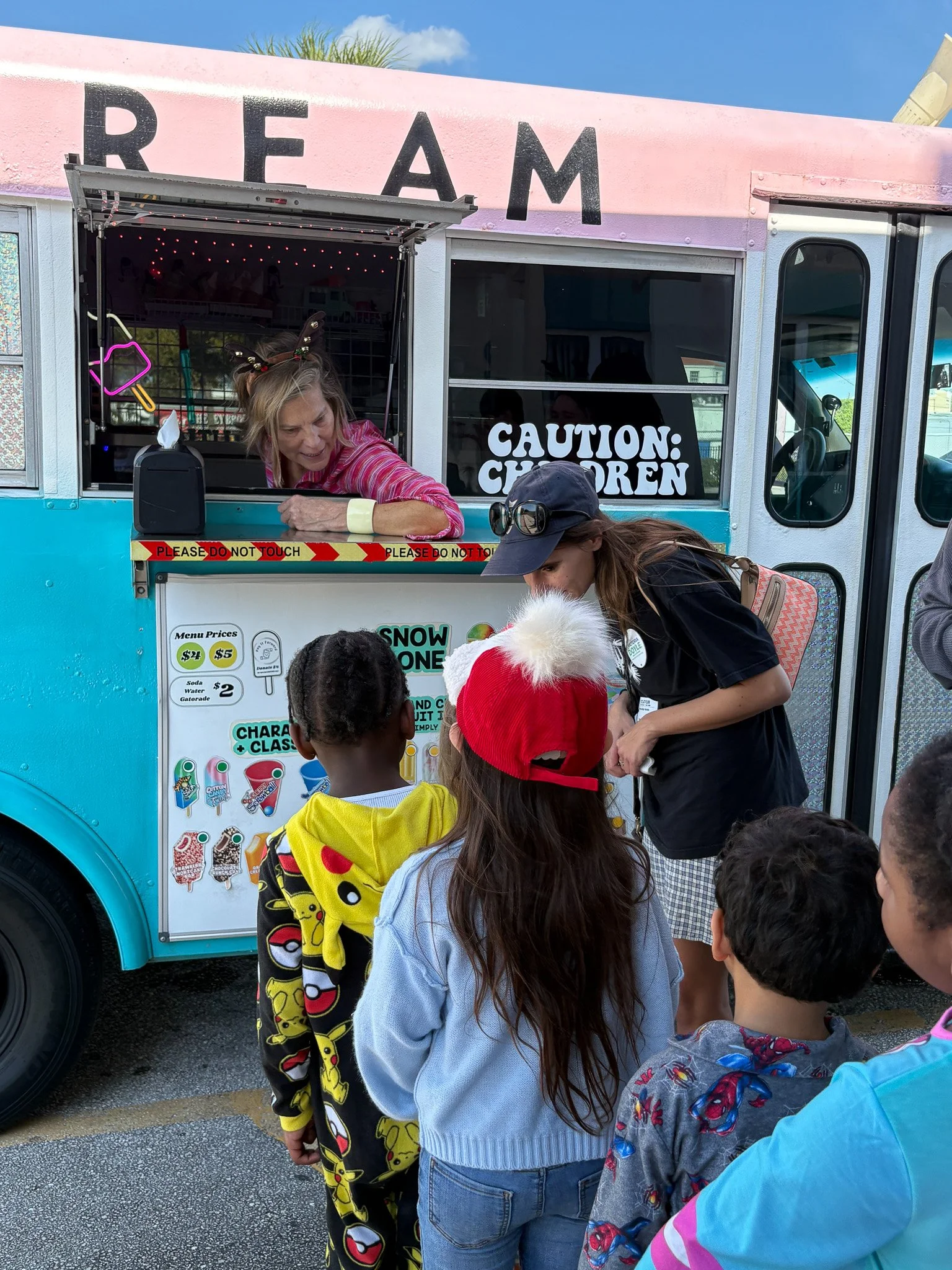 Children waiting in line at a colorful ice cream truck, with a woman working inside. The truck has a pink and blue exterior, with signage about menu prices and caution for children.