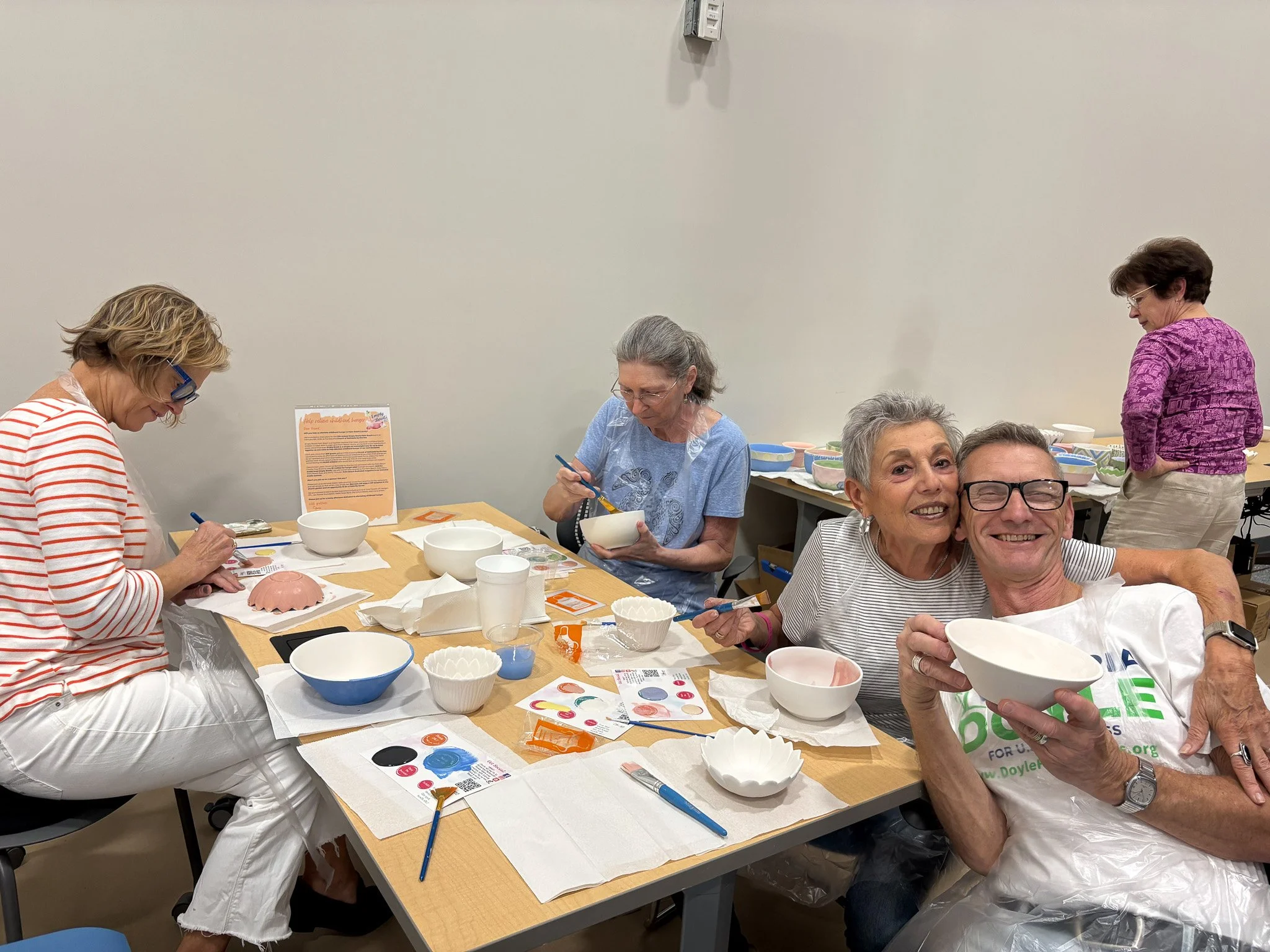 A group of five adults sitting around a table, engaging in a pottery painting activity. Two women are painting bowls, one woman is looking down and another woman stands in the background near a table with supplies. The atmosphere appears friendly and