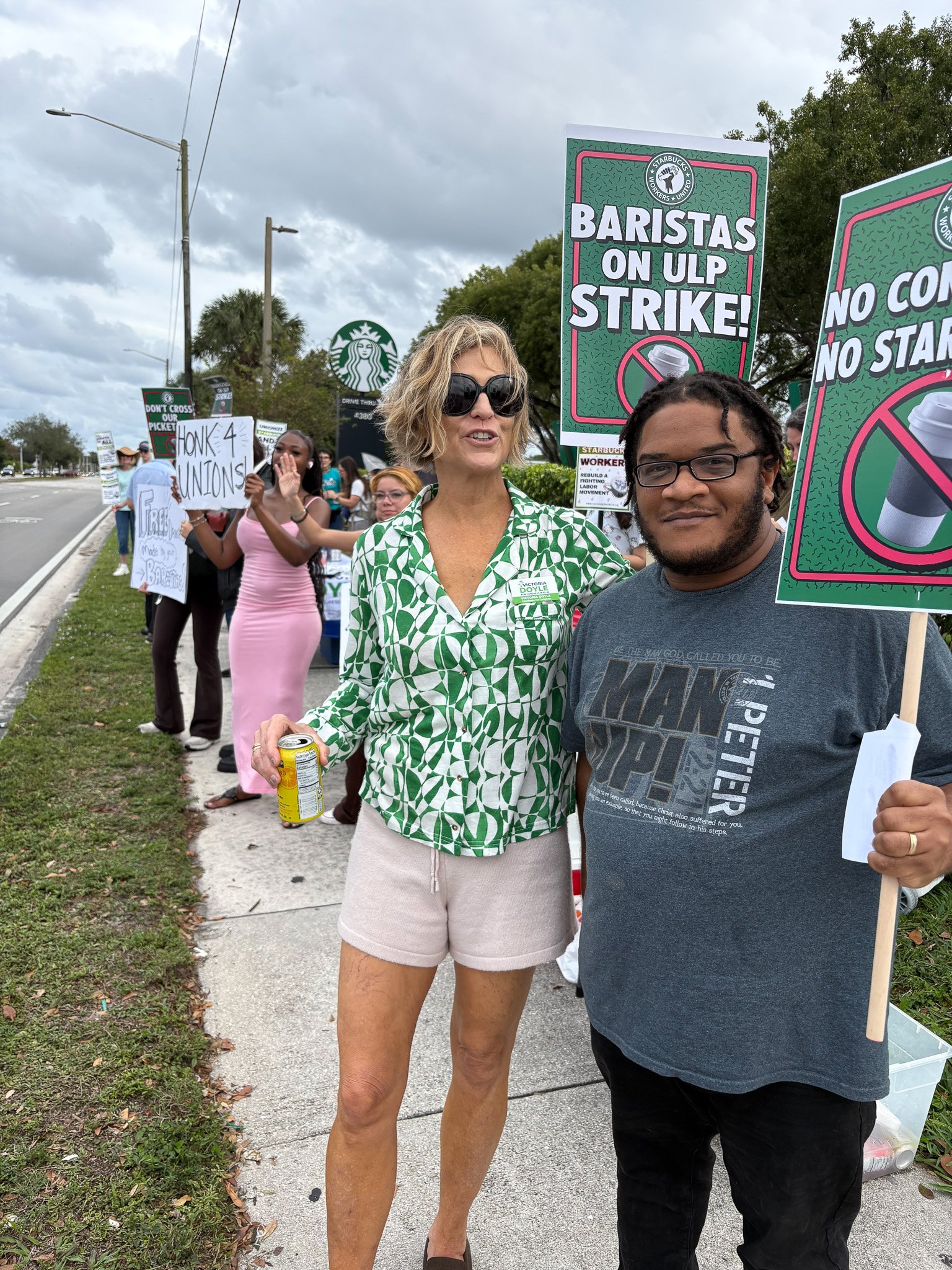 A group of protesters on a sidewalk holding signs, with a Starbucks logo visible in the background. One sign reads 'Baristas on ULP Strike!' and others contain various messages related to union and labor issues.