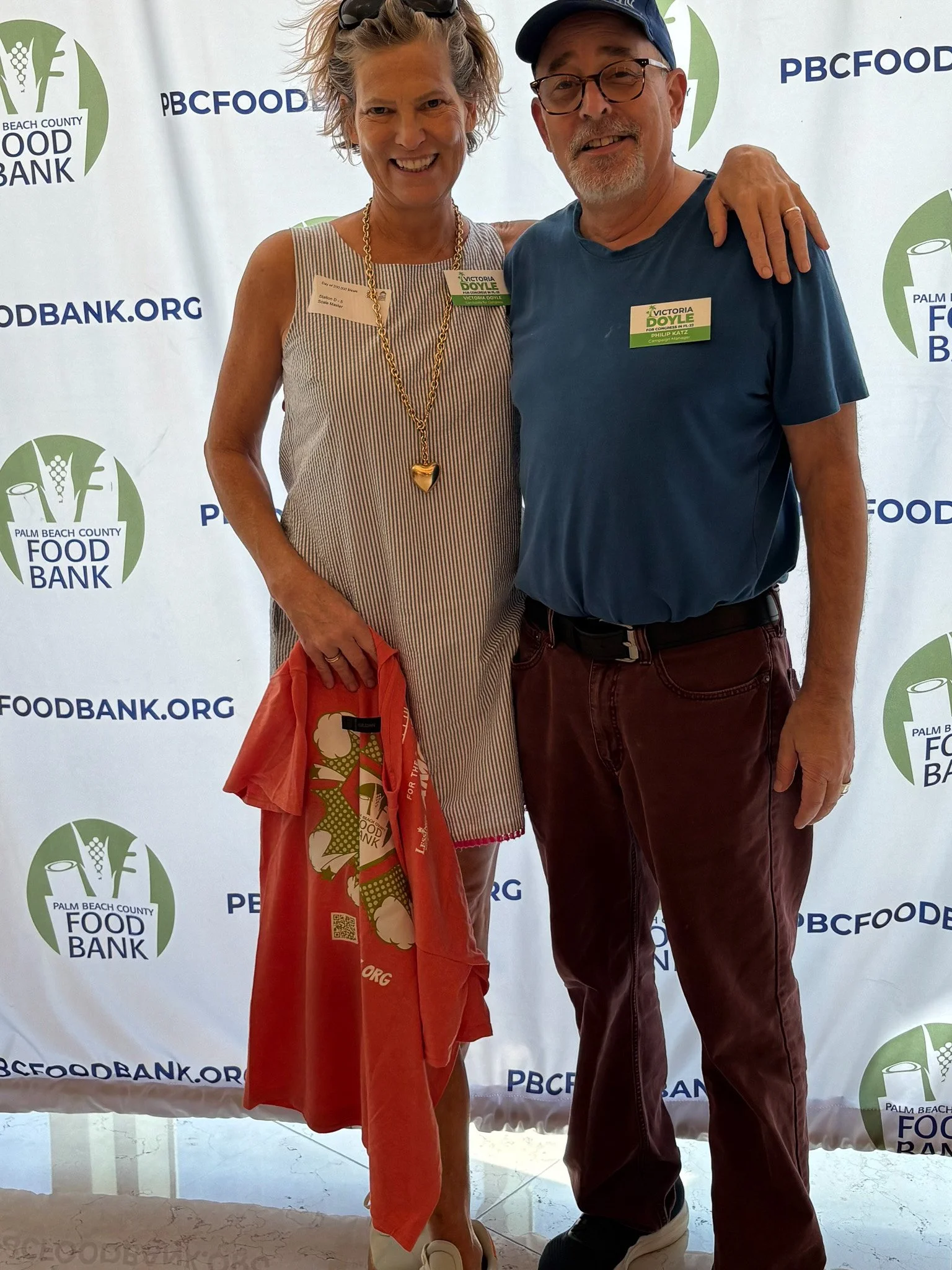 Victoria Doyle and a man smiling and standing together in front of a Palm Beach County Food Bank backdrop. The woman on the left is wearing a striped sleeveless dress, a gold necklace, and holding an orange bag. 