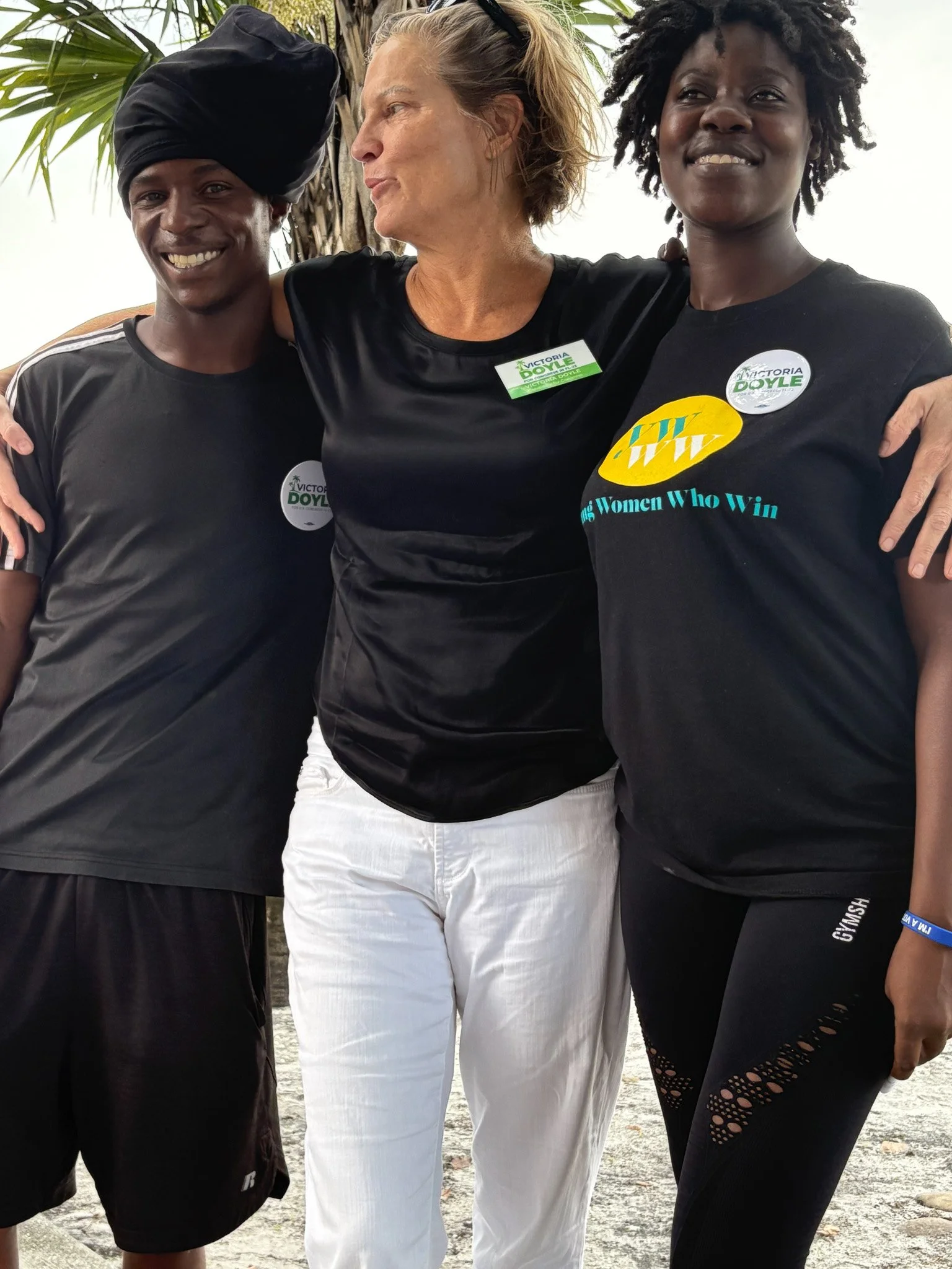 Victoria Doyle and 2 women standing together outside, embracing each other. The woman in the middle has her arms around the other two. They are smiling and wearing event badges and athletic clothing.