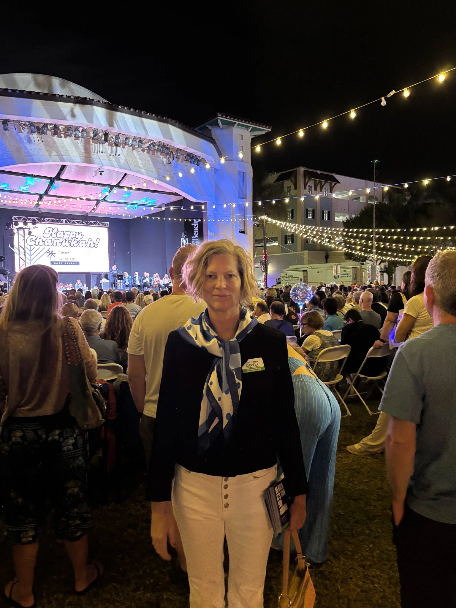 Victoria Doyle standing at an outdoor concert during the night, with a large crowd, stage with lights, and string lights overhead.