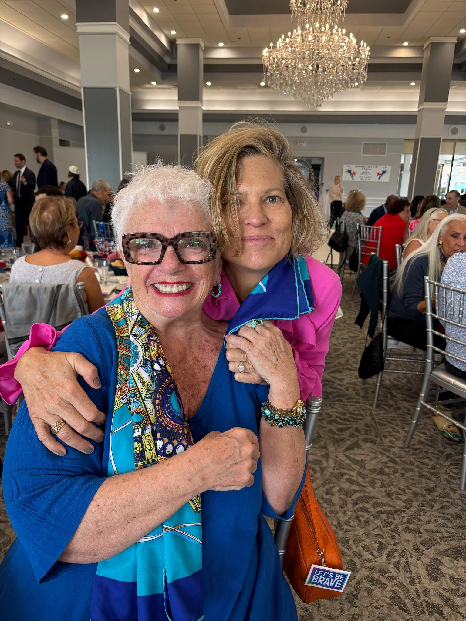 Two women hugging and smiling at a formal event in a decorated banquet hall with lots of seated guests, a chandelier, and a banner in the background. One woman has white hair, glasses, and a colorful scarf, while the other has shoulder-length blond h