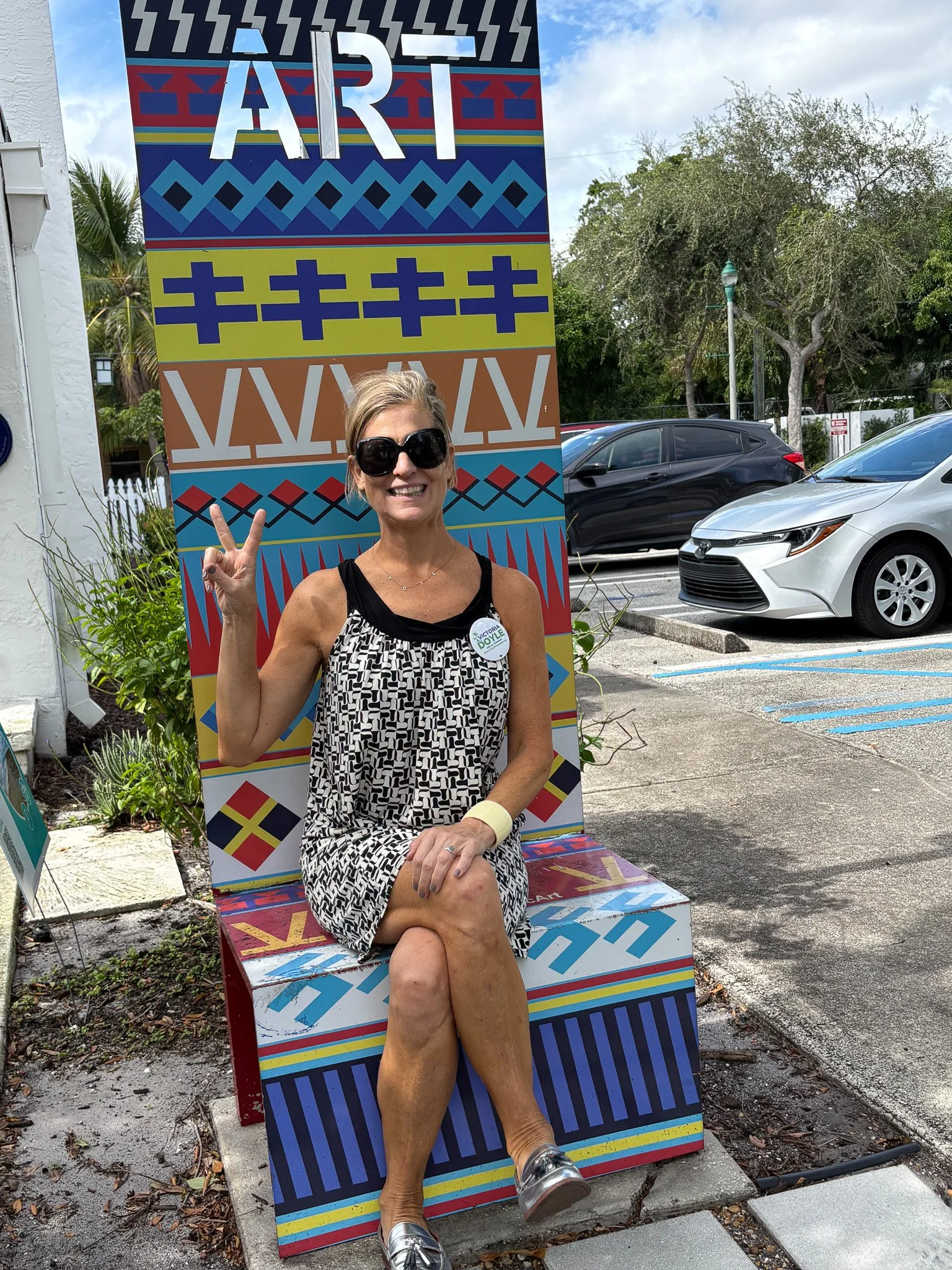 A woman with short blond hair, wearing large black sunglasses, a sleeveless black and white patterned dress, silver shoes, and a white bracelet, sitting on a colorful, patterned art installation bench outside, making a peace sign with her hand, smili