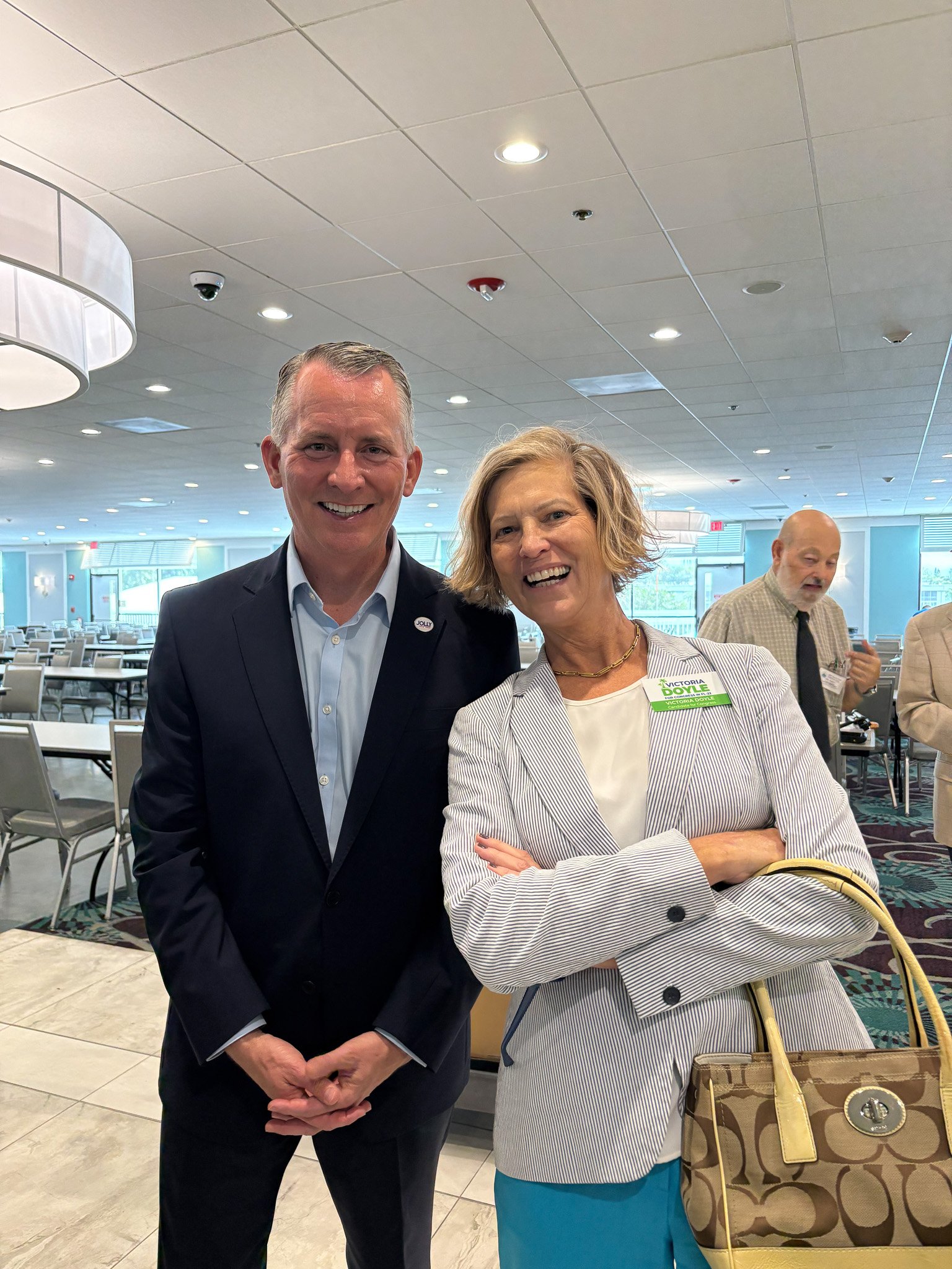 A man and a Victoria Doyle standing in a conference room, smiling at the camera. The man is wearing a dark suit and light blue shirt, and the woman is wearing a light-colored blazer and blue pants, carrying a patterned handbag.
