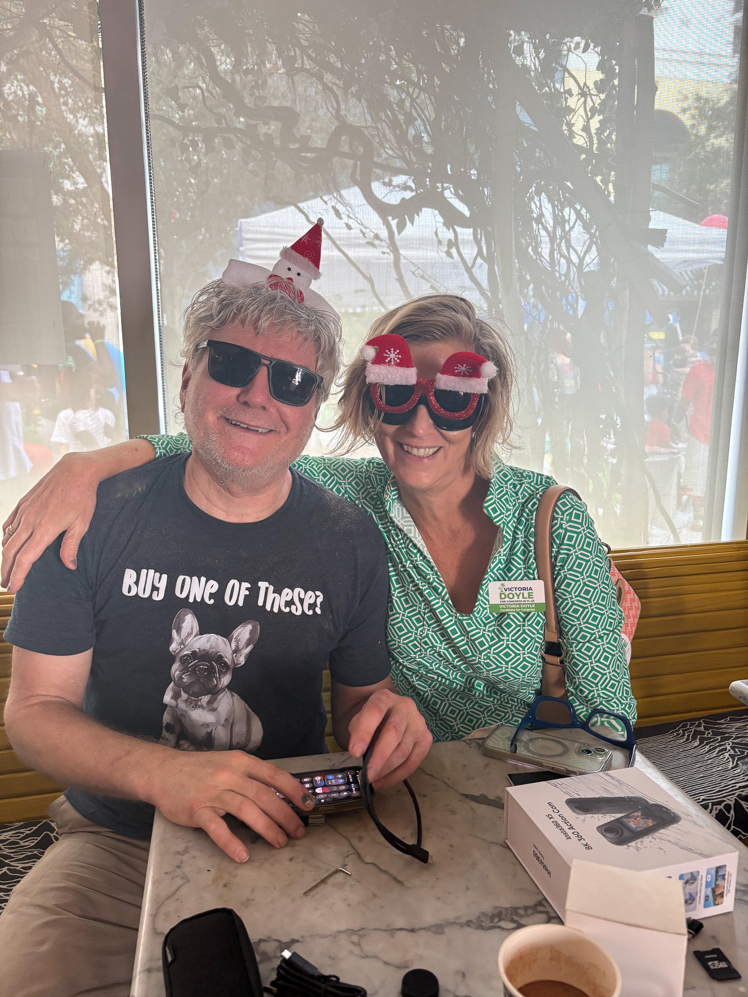 Two people sitting at a table wearing holiday-themed sunglasses and accessories. The man has a small Santa hat on his head and dark sunglasses, and the woman has glasses with Christmas hats on them. They are smiling, and the man is holding a phone. T