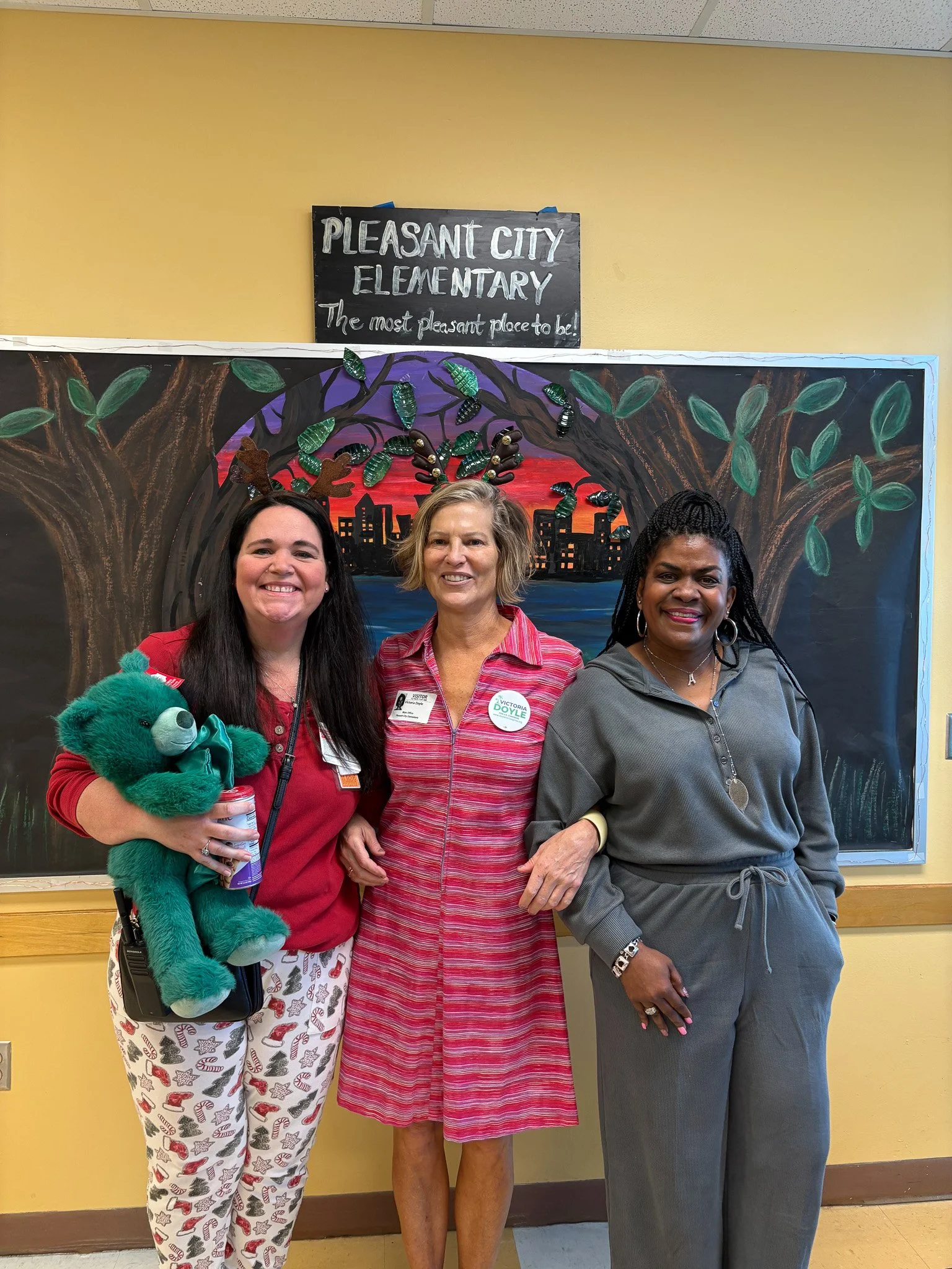 Three women standing in front of a blackboard with a colorful background painting of a cityscape under a sunset, celebrating Pleasant City Elementary. The women are smiling, and one is holding a green teddy bear.Inside classwroom
