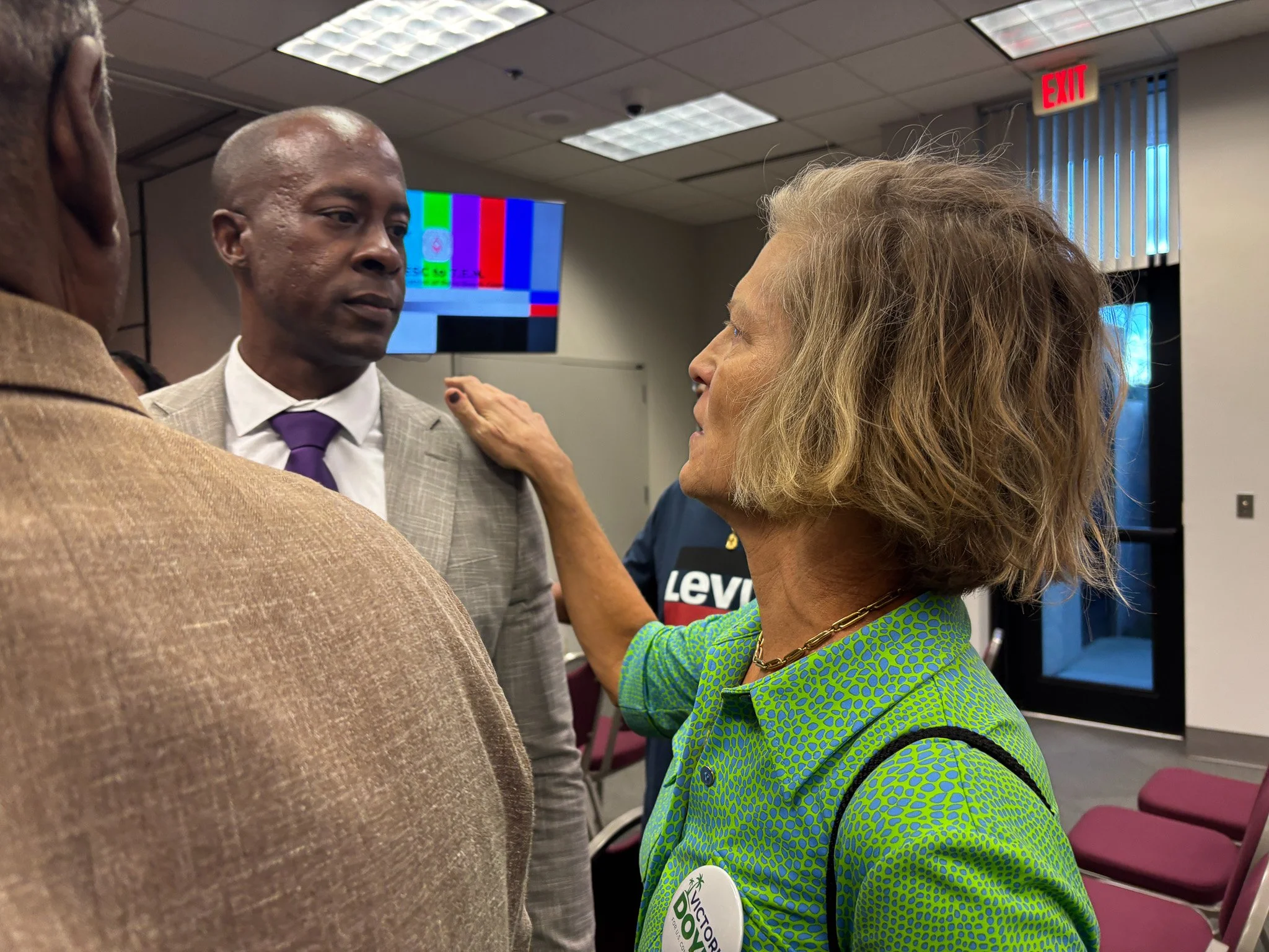 Victoria Doyle, A woman with short curly blonde hair and wearing a green patterned shirt is touching the shoulder of a man in a gray suit and purple tie. They are in a room with purple chairs and a TV displaying color test patterns.