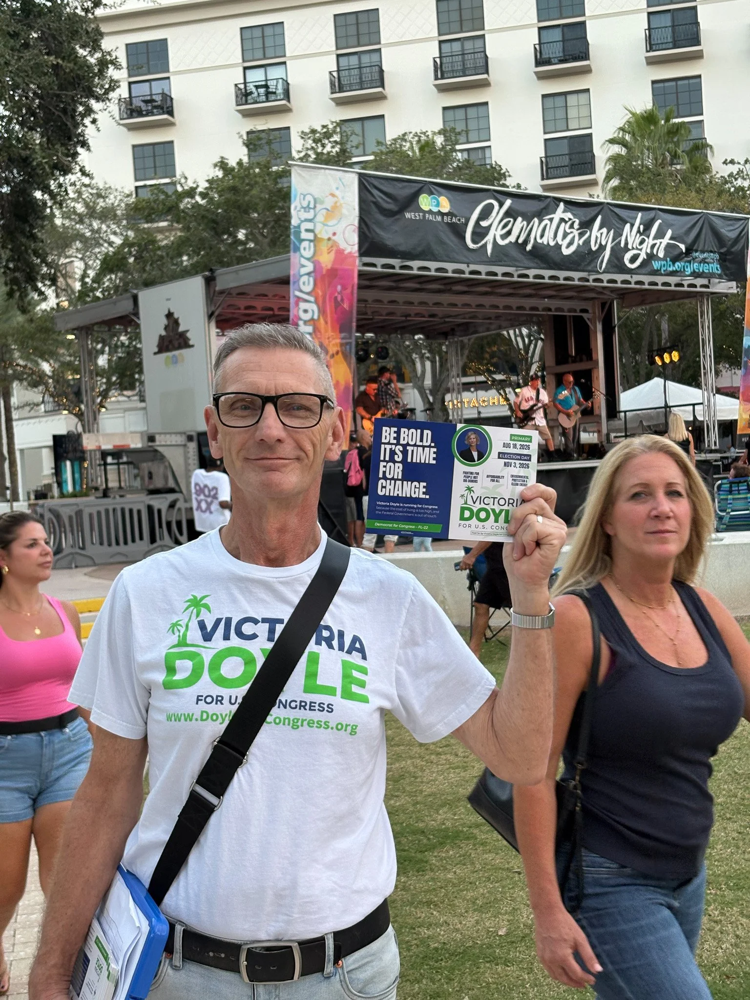 Man holding a campaign sign at an outdoor event, wearing a white T-shirt supporting Victoria Doyle for U.S. Congress, with stage and people in the background.