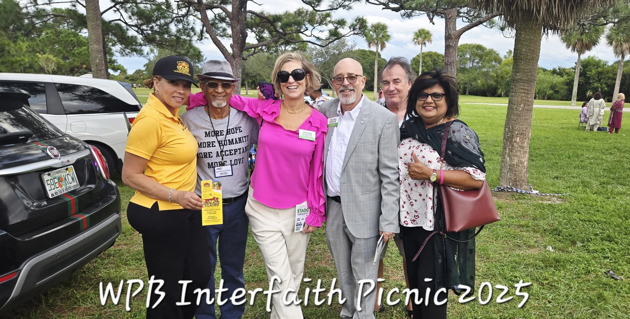 Six people standing together at WPB Interfaith Picnic 2023, outdoors on a grassy area with trees and parked cars in the background. They are smiling and dressed casually or semi-formally.