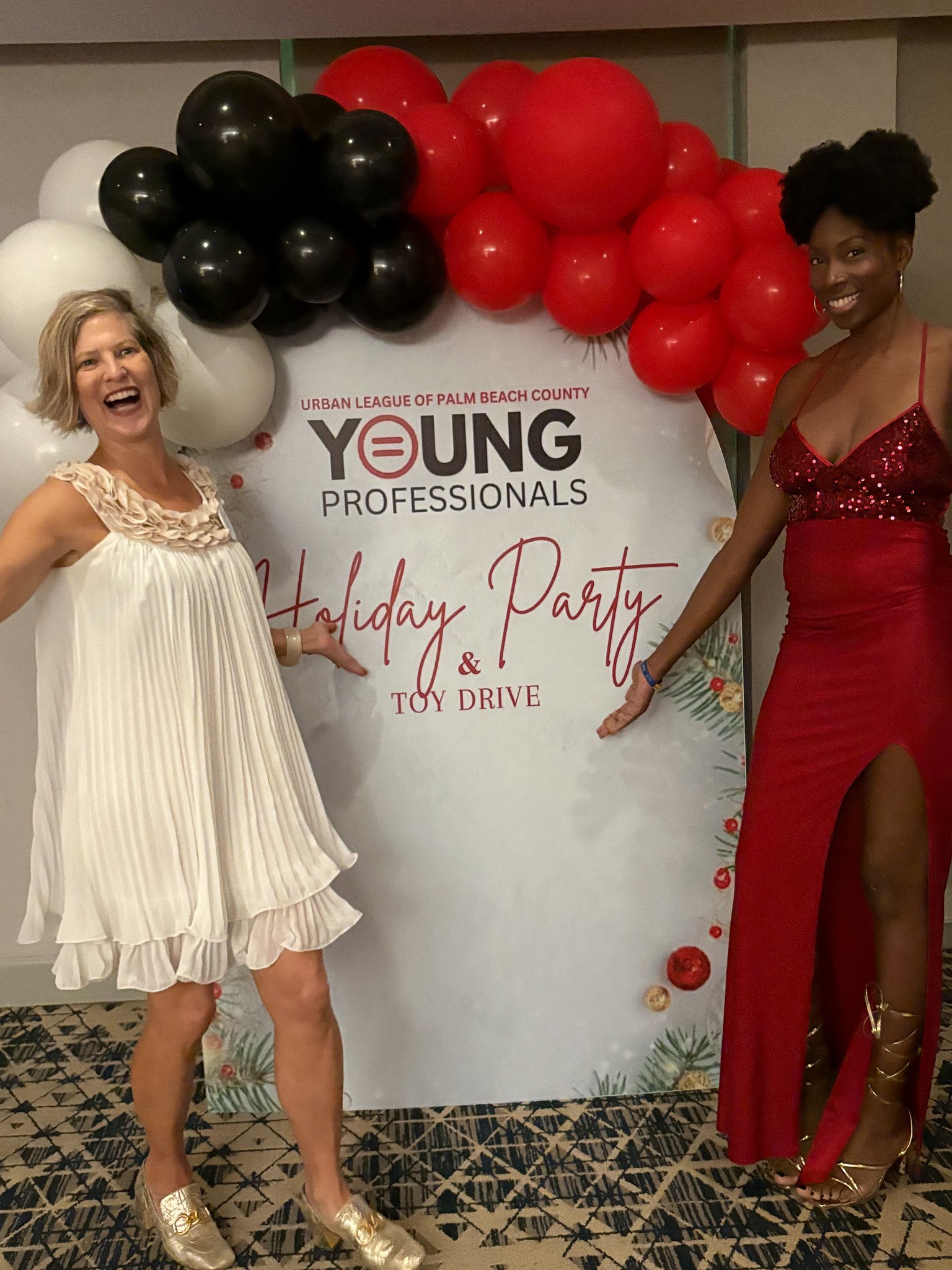 Two women posing and smiling at a holiday party and toy drive event, standing next to a white sign decorated with balloons and holiday ornaments.