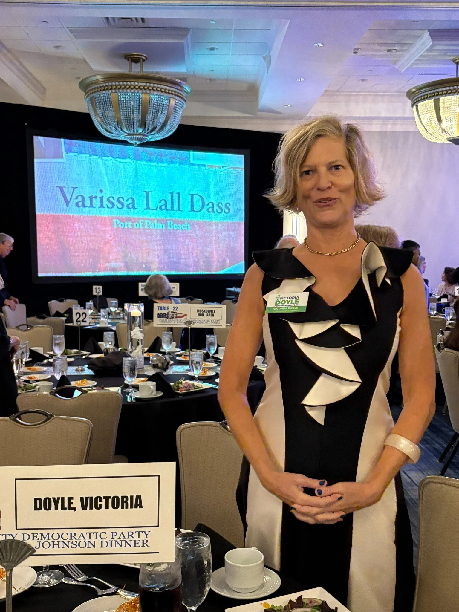 Victoria Doyle standing at a banquet table during a formal event with a large screen behind her displaying her name, 'Varissa Lall Dass,' and the location, 'Port of Palm Beach.' 