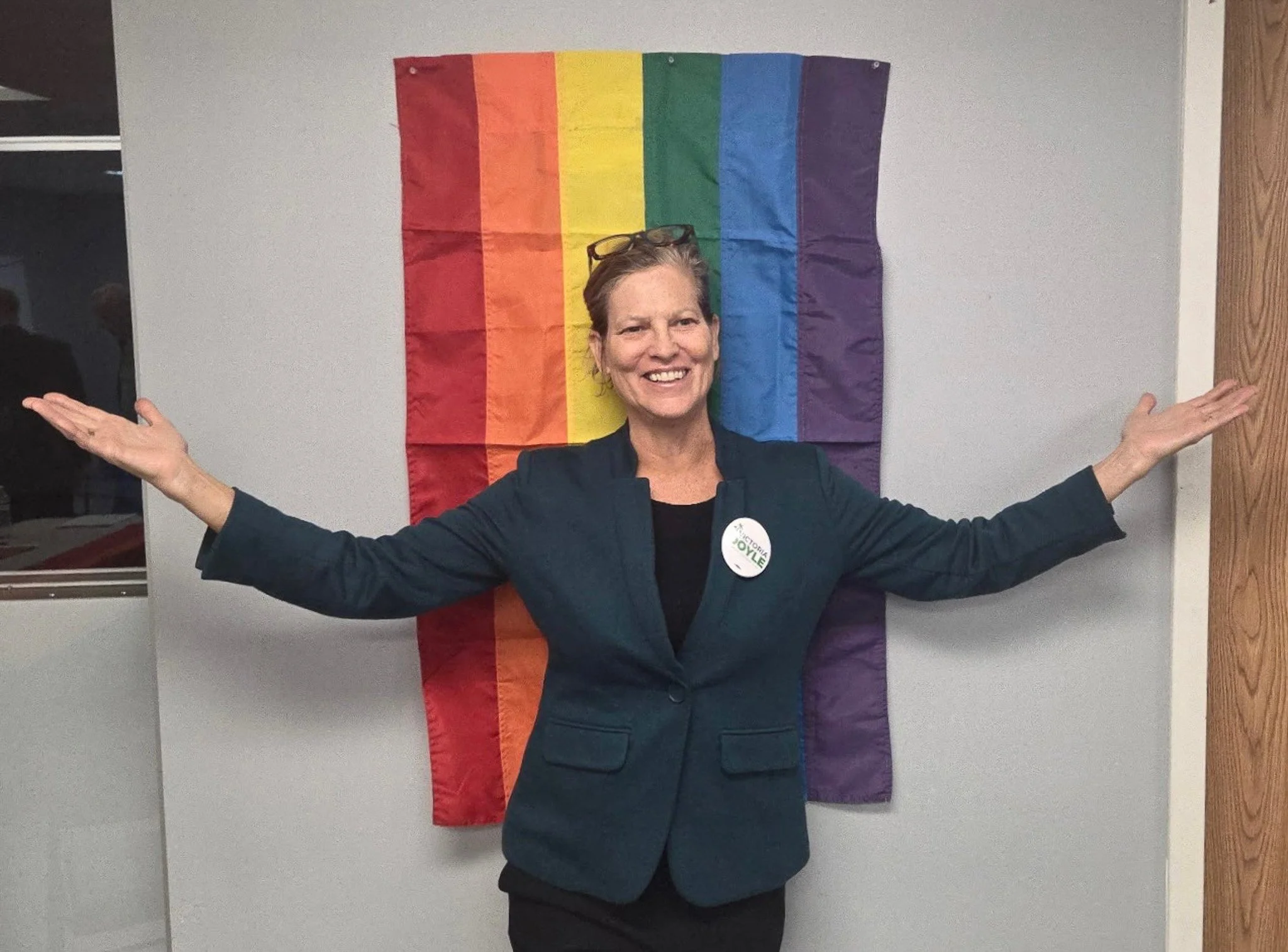 Victoria Doyle standing in front of a rainbow pride flag with her arms outstretched. She is smiling and wearing a dark blazer with a campaign button. The background includes a wall and a window.