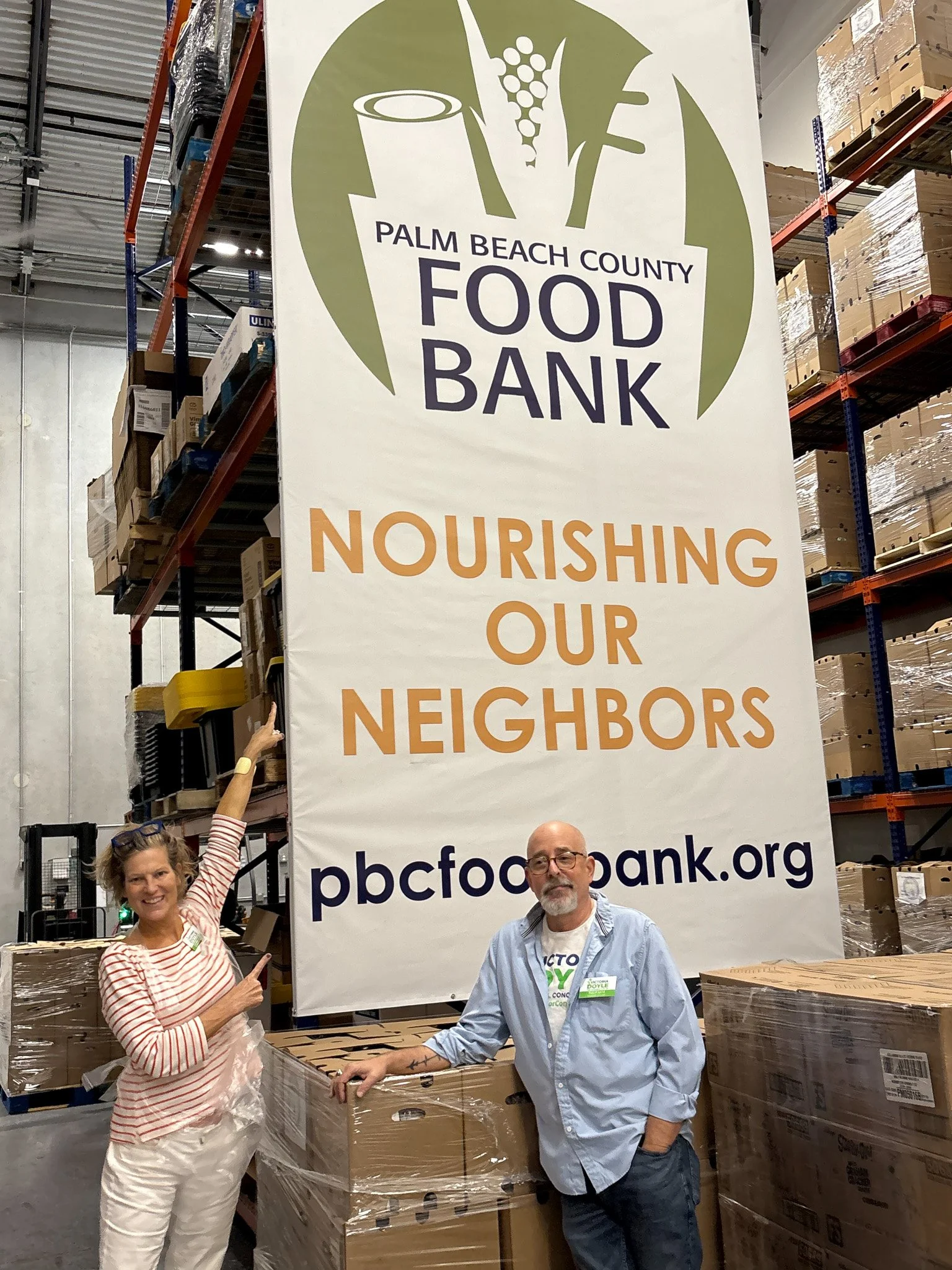Victoria Doyle and a man standing inside a warehouse in front of a large Palm Beach County Food Bank banner that reads 'Nourishing Our Neighbors'. They are surrounded by stacked boxes for food distribution.