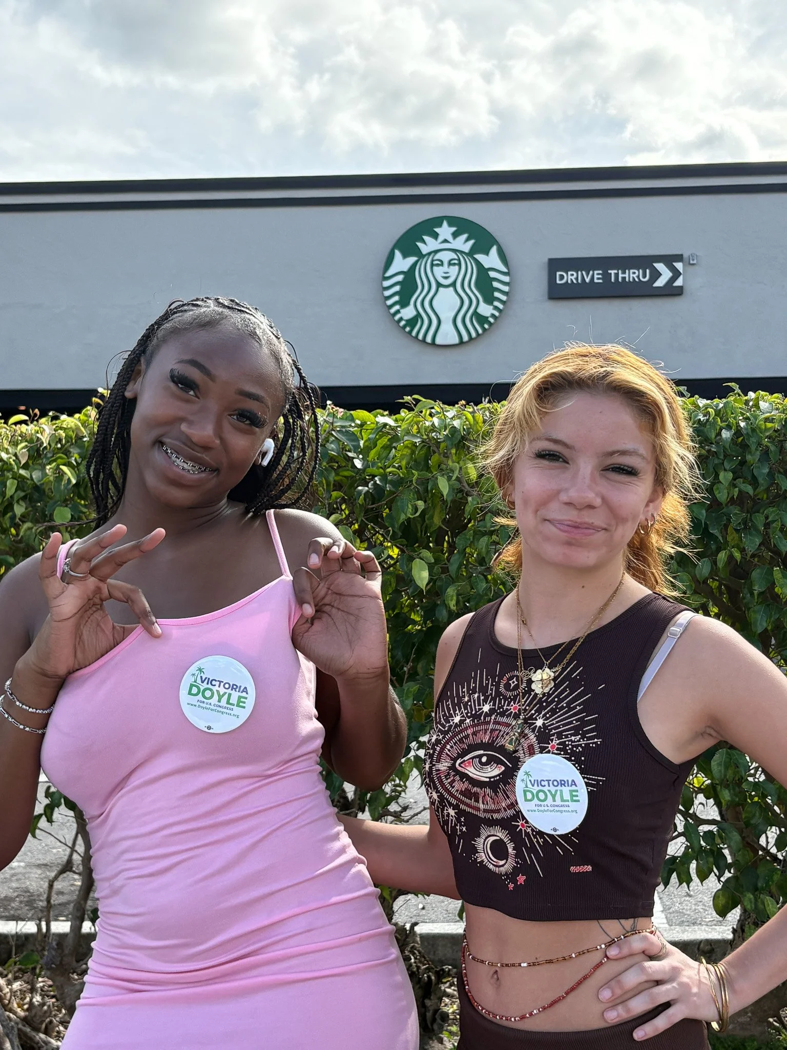 Two young women standing outdoors in front of a hedge and a Starbucks store, smiling at the camera. They are wearing campaign buttons that say "Victoria Doyle for Congress."
