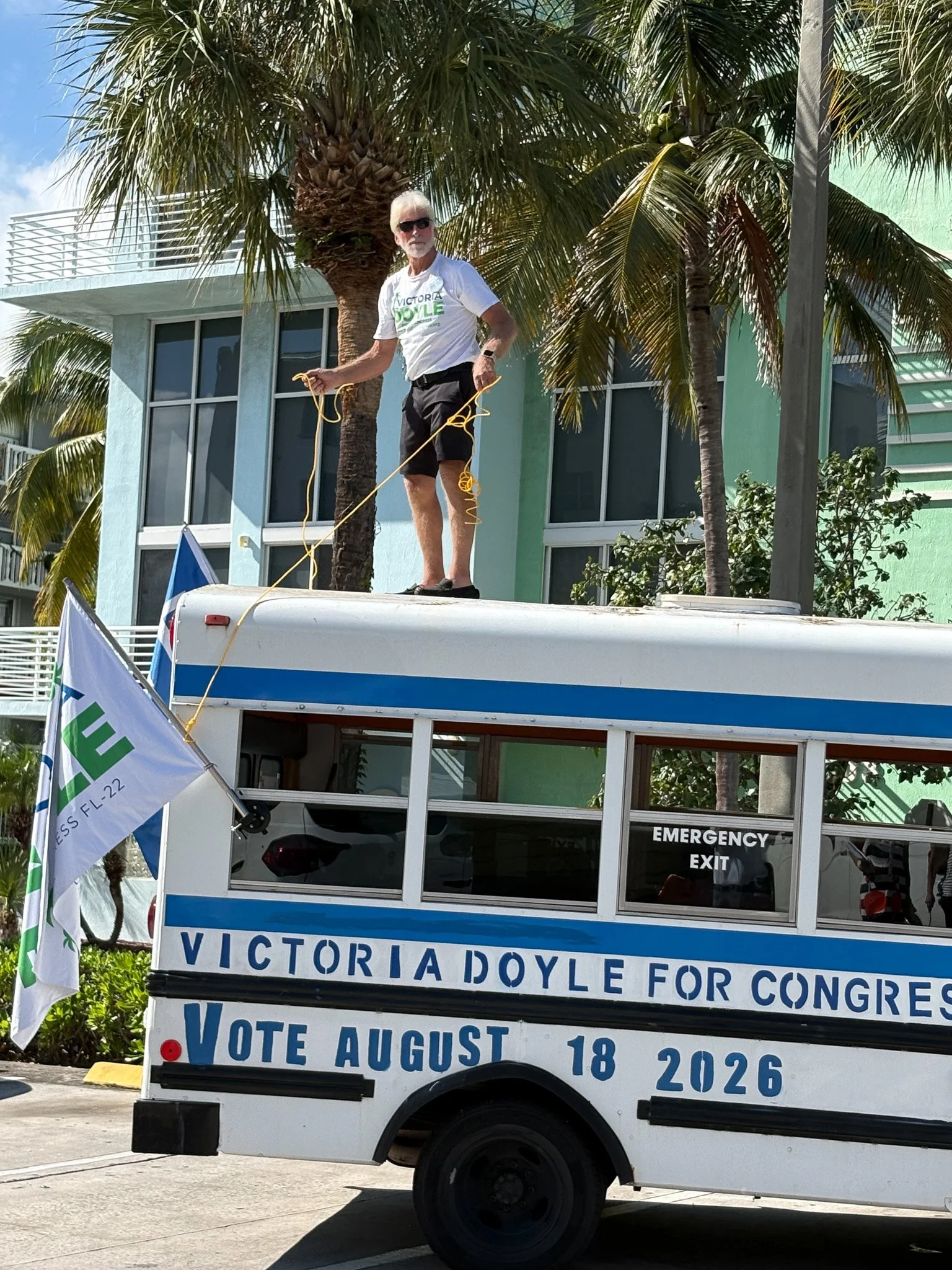 A man standing on top of a campaign bus for Victoria Doyle, holding a yellow rope, with palm trees and a colorful building in the background. The bus has campaign signage including the message 'Victoria Doyle for Congress' and 'Vote August 18, 2026'.