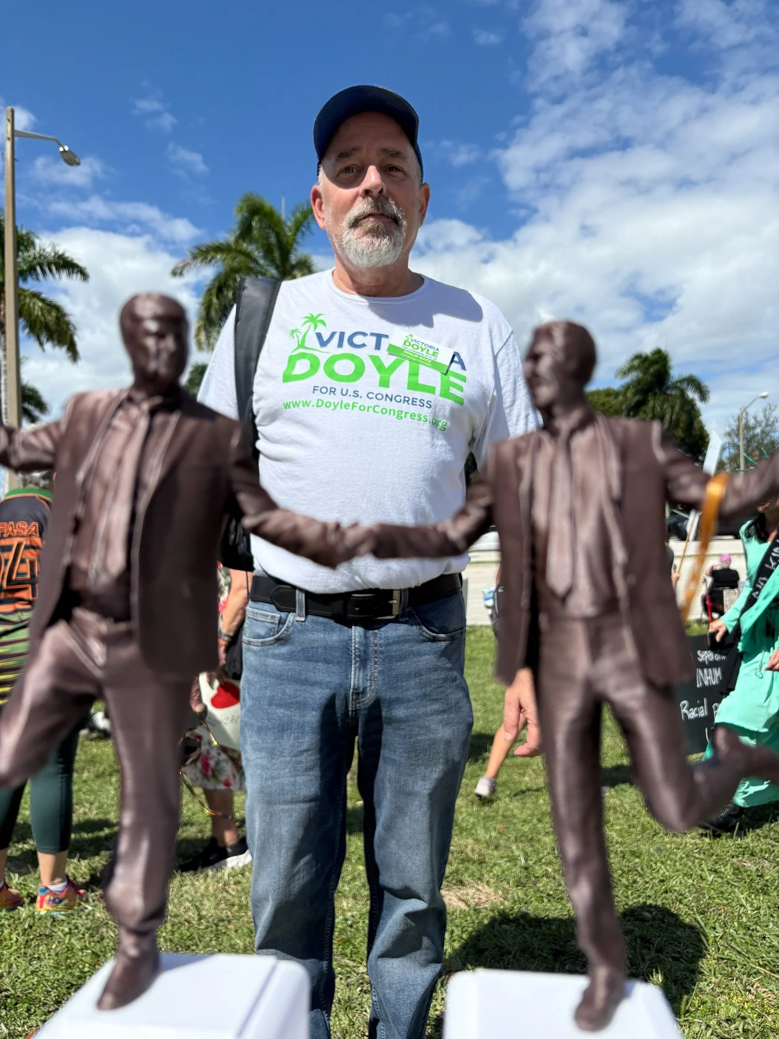 Philip Katz wearing a white T-shirt with green and blue campaign text, standing outdoors among palm trees, with a decorative bronze sculpture of two men. Clouds and blue sky in the background.