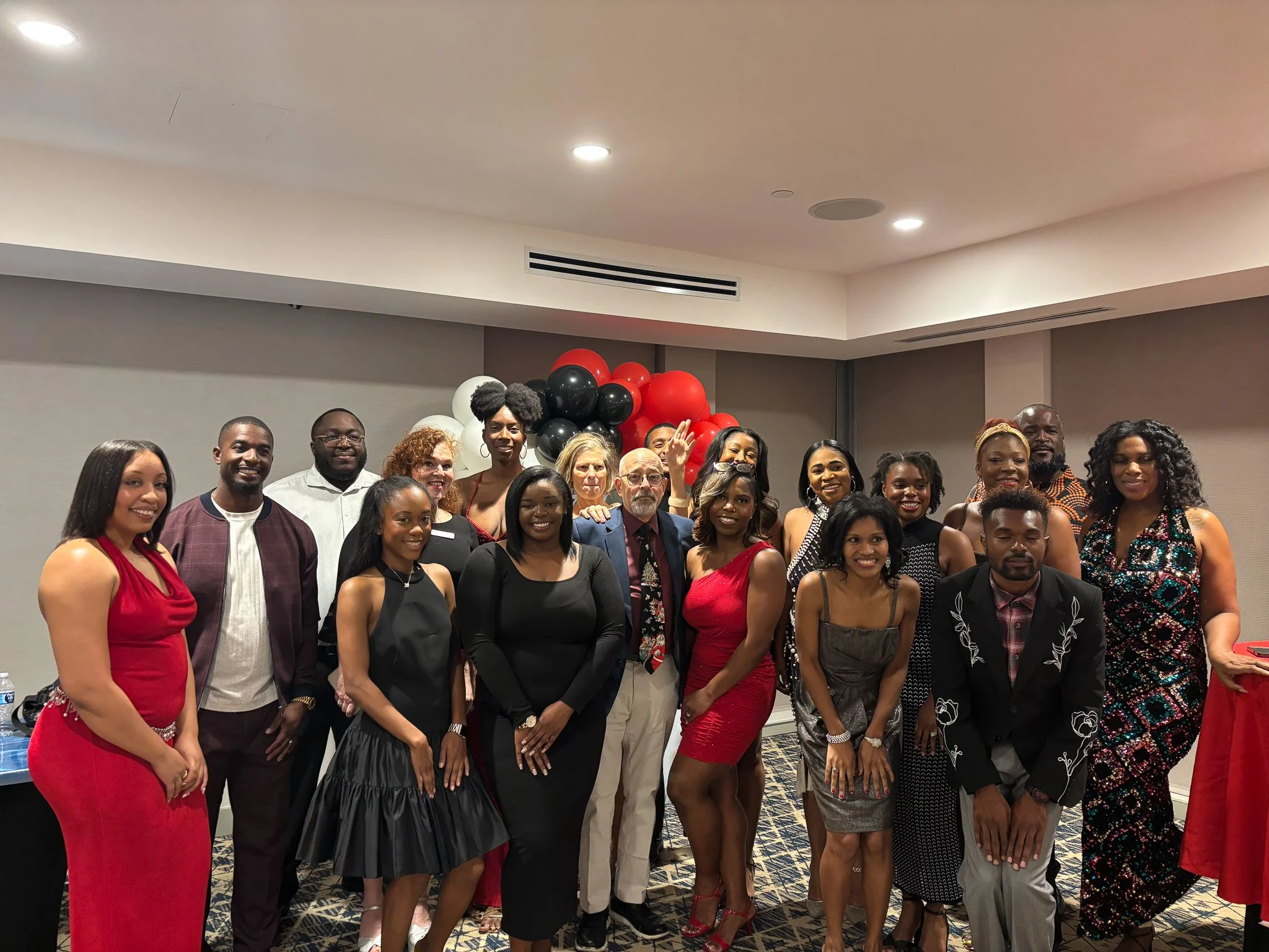 Group of people dressed in formal attire standing together indoors in front of a decorated wall with red, black, and white balloons.