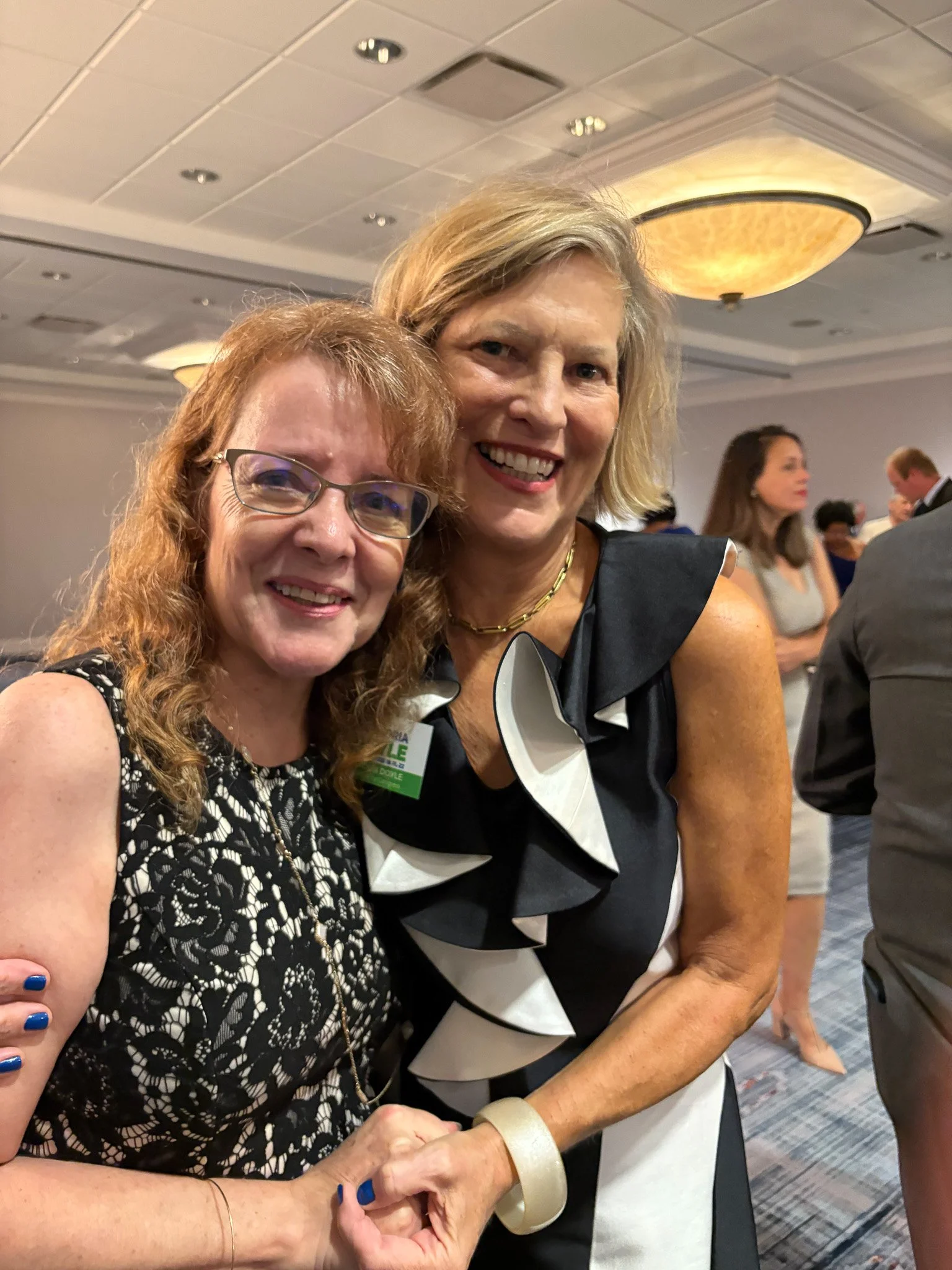 Two women smiling and posing together at an indoor event, with people and tables in the background.