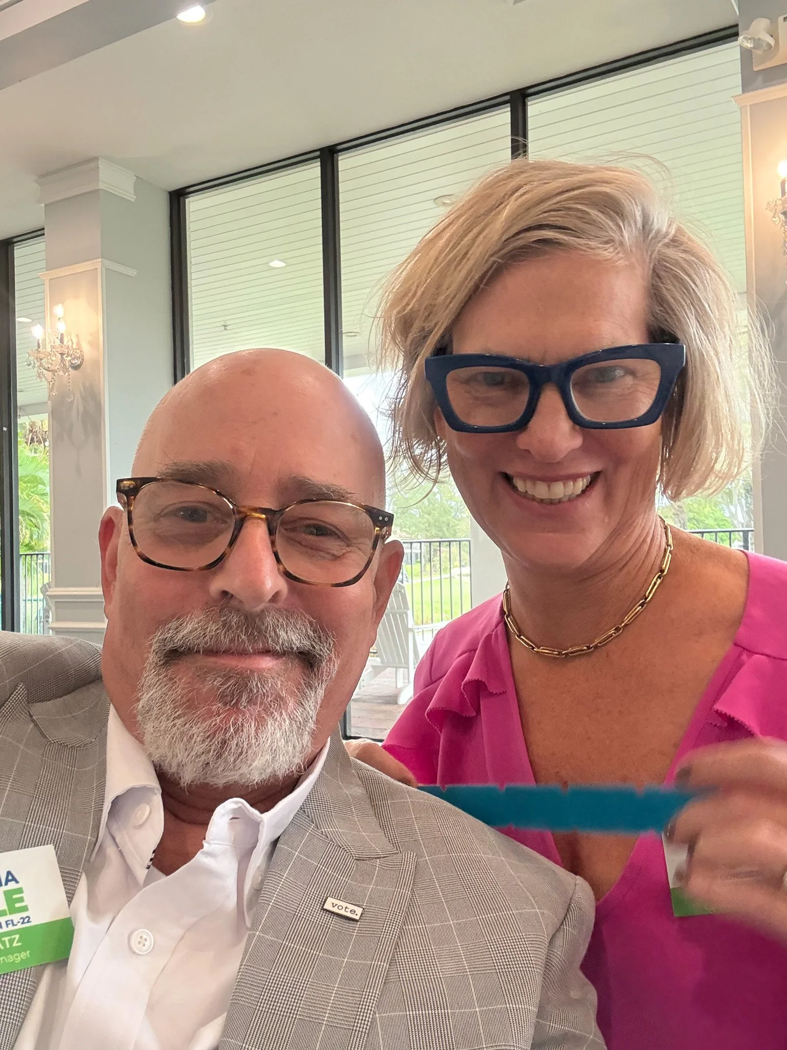 Victoria Doyle and Philip Katz - A smiling man with glasses and a beard sits next to a smiling woman with glasses and a gold chain necklace, both wearing name tags, indoors with large windows and outdoor greenery visible in the background.