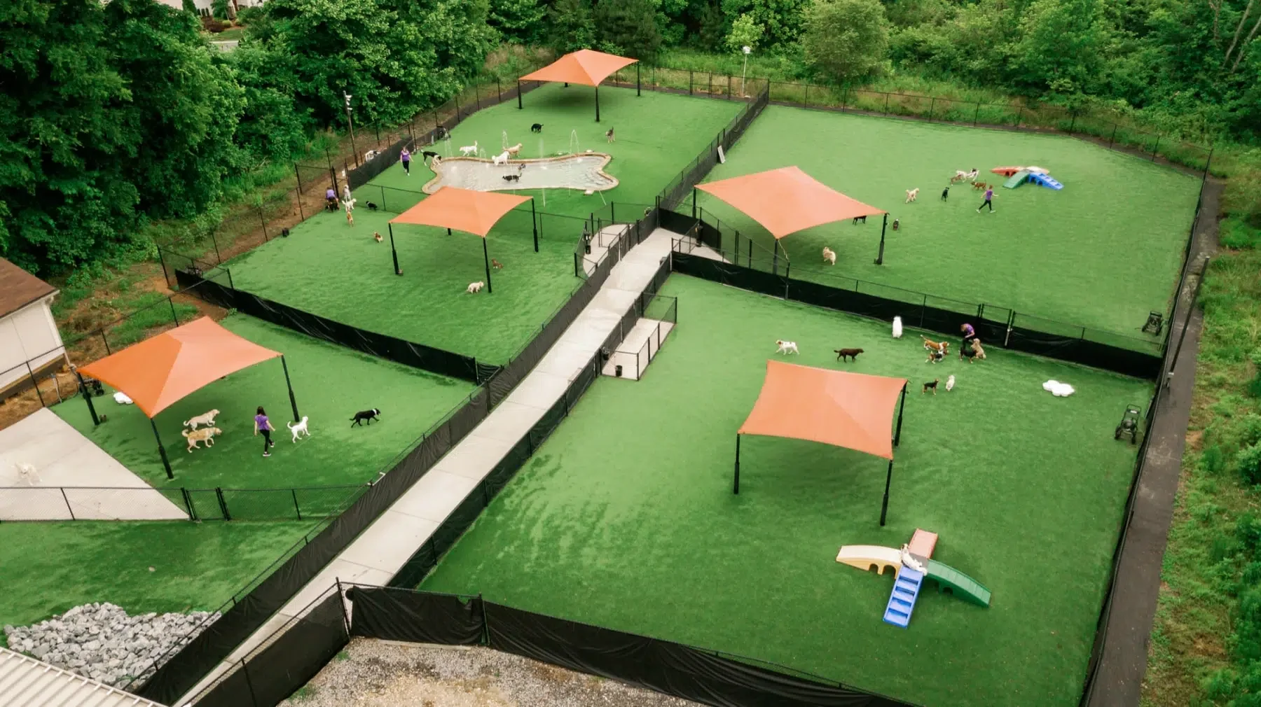 Aerial view of an outdoor dog park with four separate grassy fenced areas, each with shade structures, dog equipment, and several dogs playing, surrounded by trees.