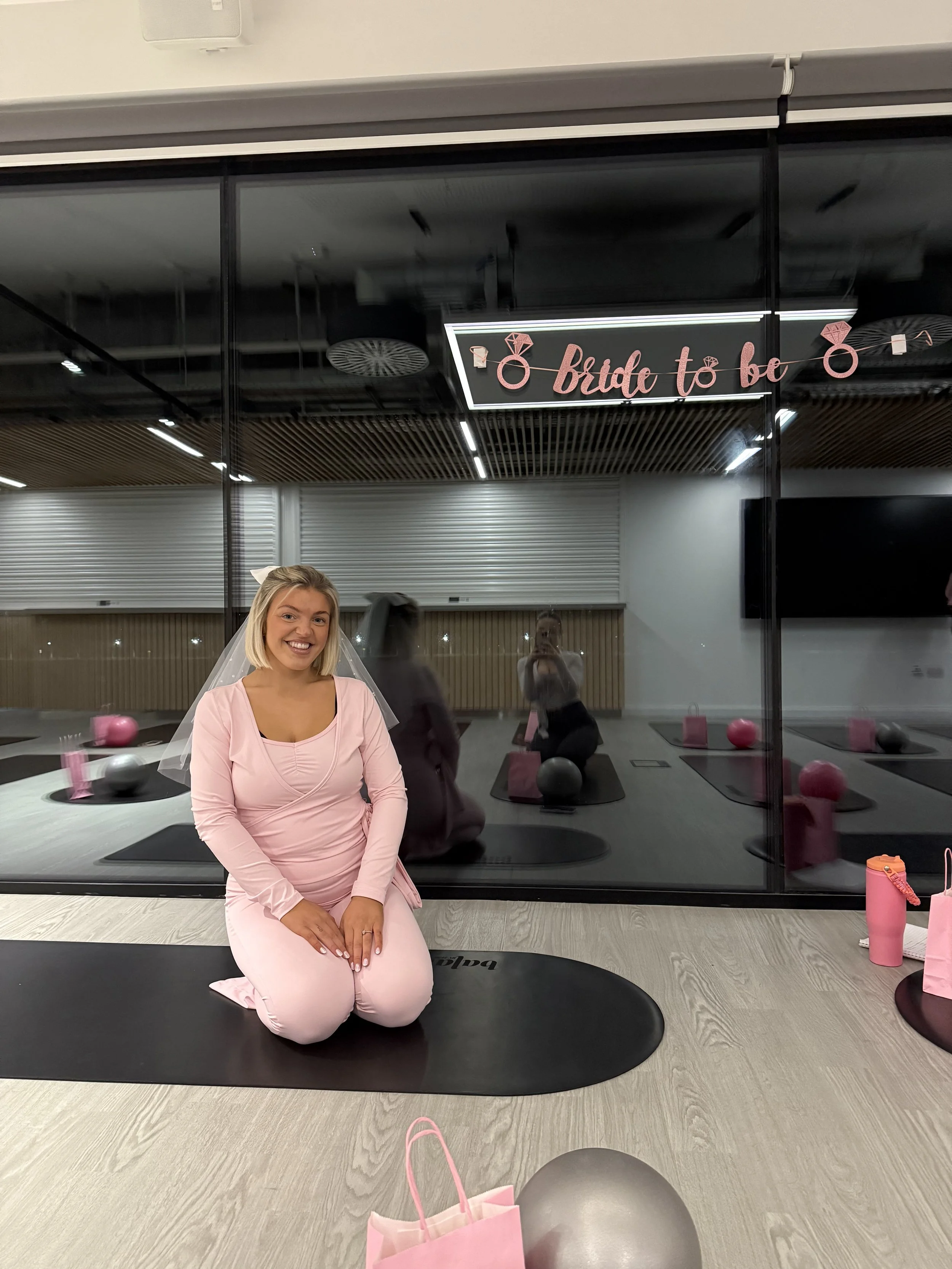 A woman dressed in pink, kneeling on a black exercise mat, smiling at the camera inside a fitness studio with pink and gray decorations, including a sign that reads 'bride to be' and a pink gift bag in the foreground.
