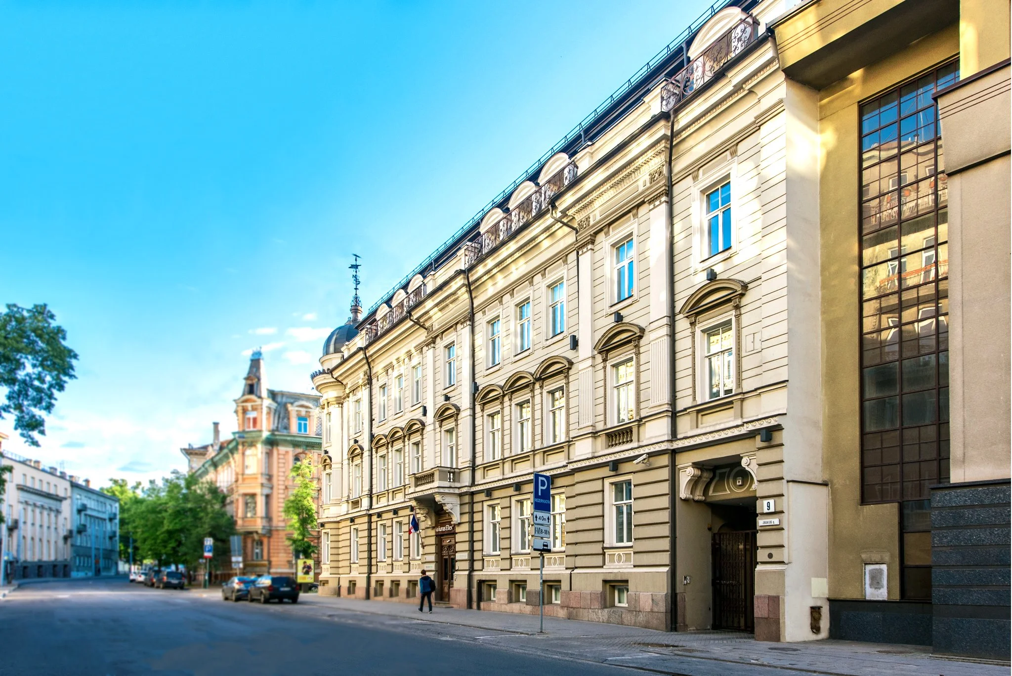 Istorinio europietiško stiliaus pastato vaizdas iš gatvės – su puošnia architektūra, langais ir stovėjimo ženklu, po mėlynu dangumi.