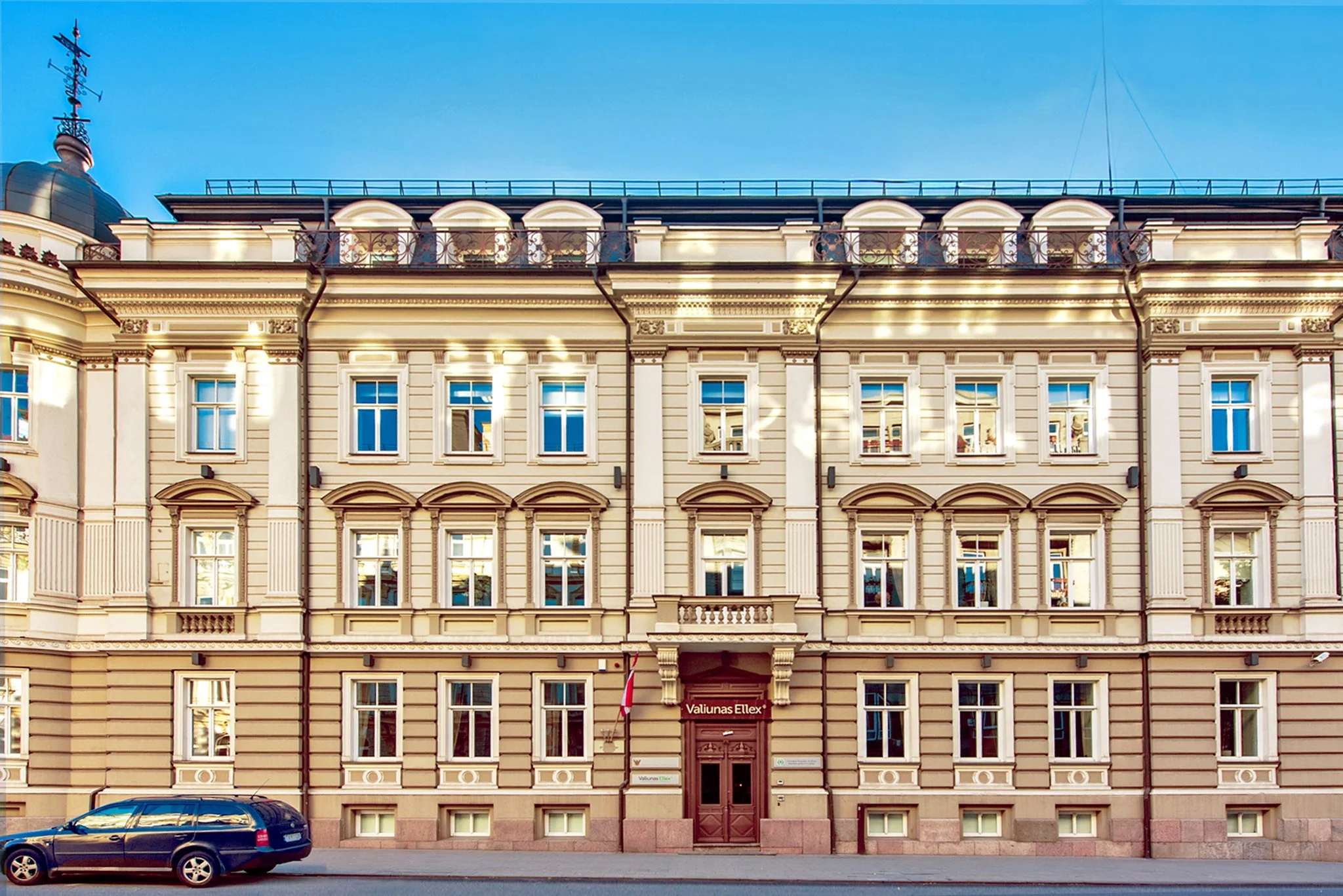 Istorinis smėlio spalvos pastatas su dekoruotais langų rėmais, kolonomis ir balkonais, priekyje pastatytas juodas automobilis.