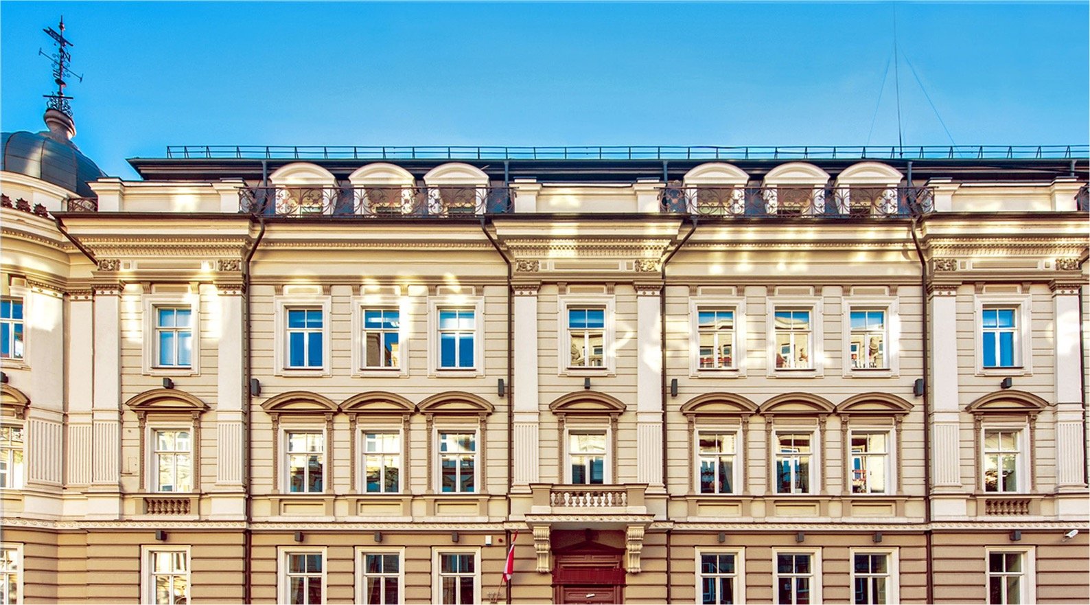 Istorinis smėlio spalvos pastatas su puošniomis architektūrinėmis detalėmis ir daugybe langų, po giedru mėlynu dangumi.