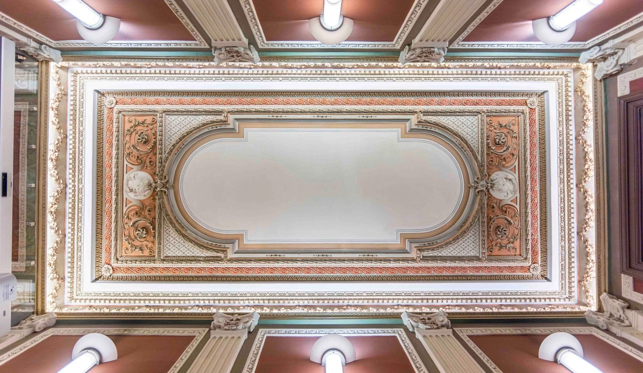 Istorinės patalpos lubos su puošniais lipdiniais, dekoratyviniais elementais ir apšvietimo šviestuvais.