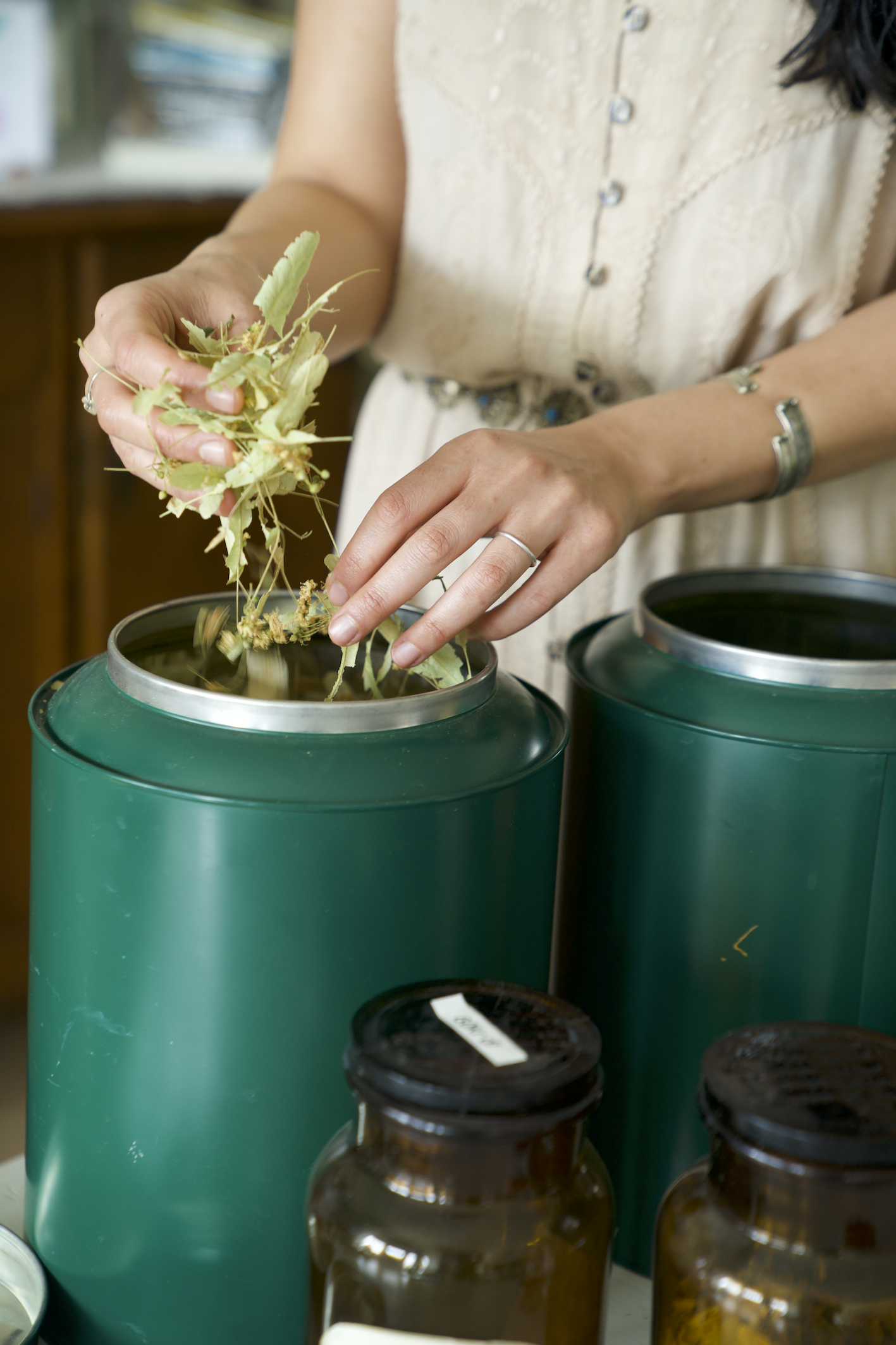 Person using a green herbal medicine or tea infuser, adding dried herbs or plant material into a green container, with brown glass jars nearby.