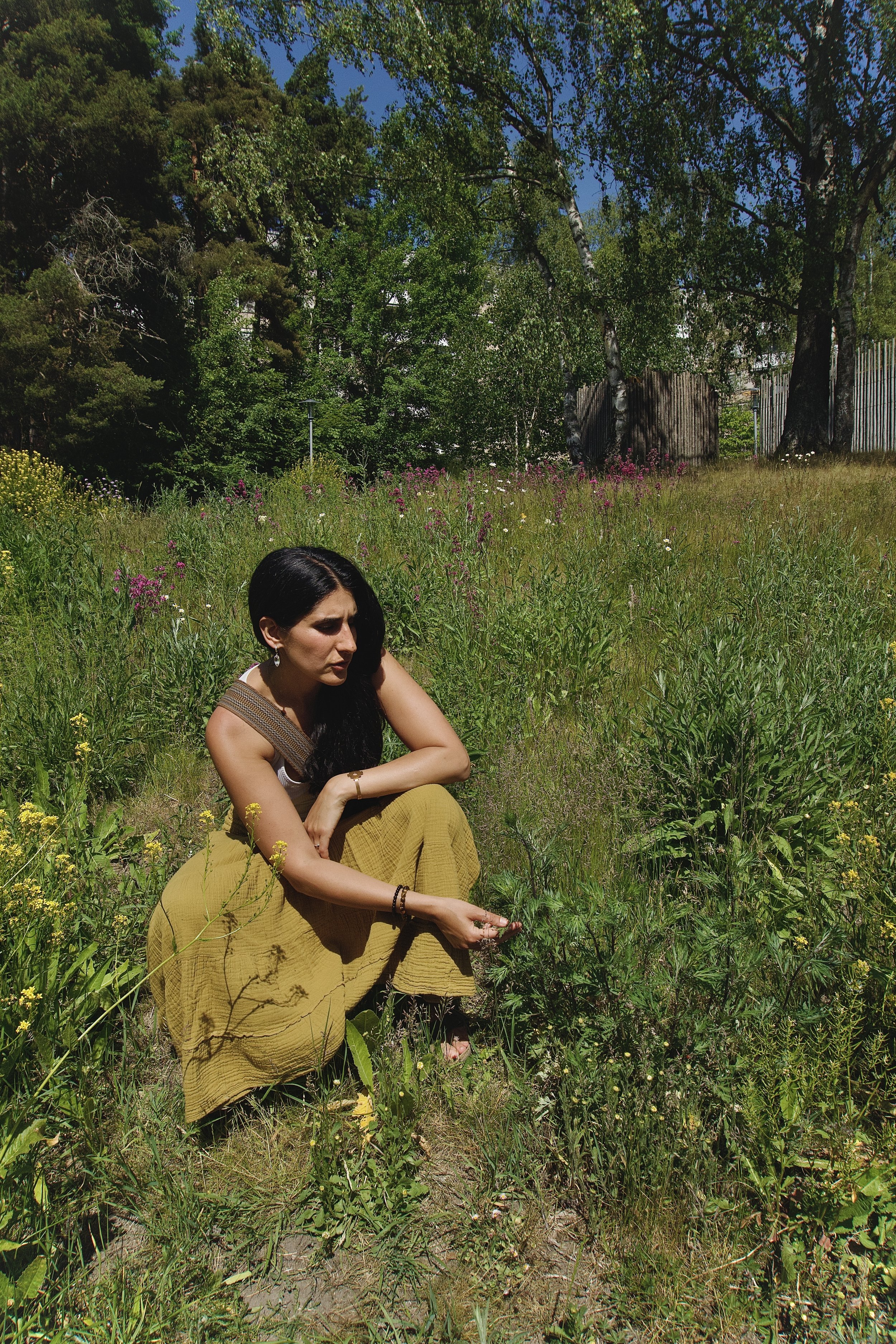 A woman with dark hair, dressed in a yellow skirt and sleeveless top, crouched in a field of wildflowers and greenery, with trees and a fence in the background, under a bright blue sky.