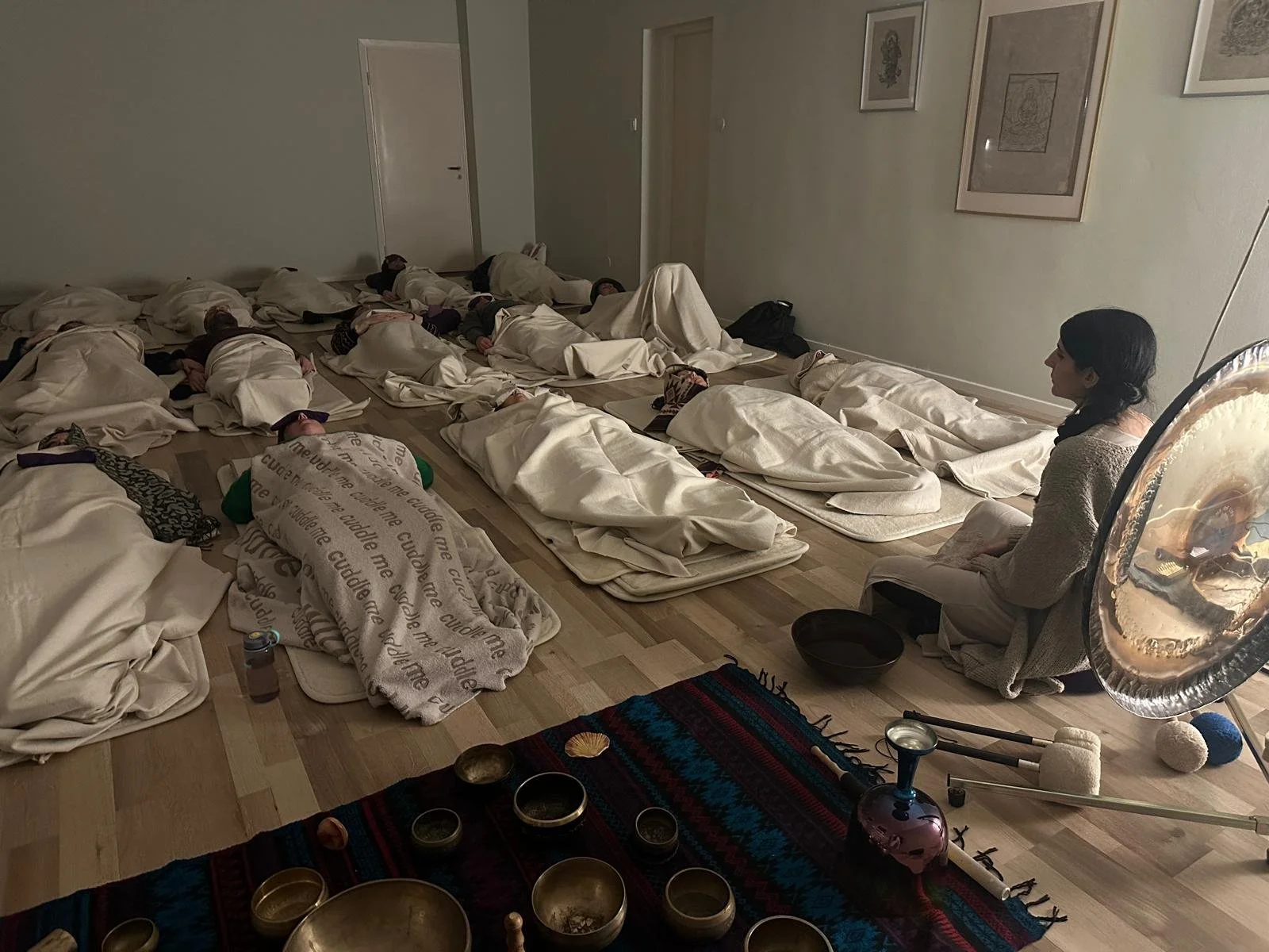 A group of people practicing meditation in a dimly lit room, laying on mats covered with blankets, with a woman sitting cross-legged and observing.