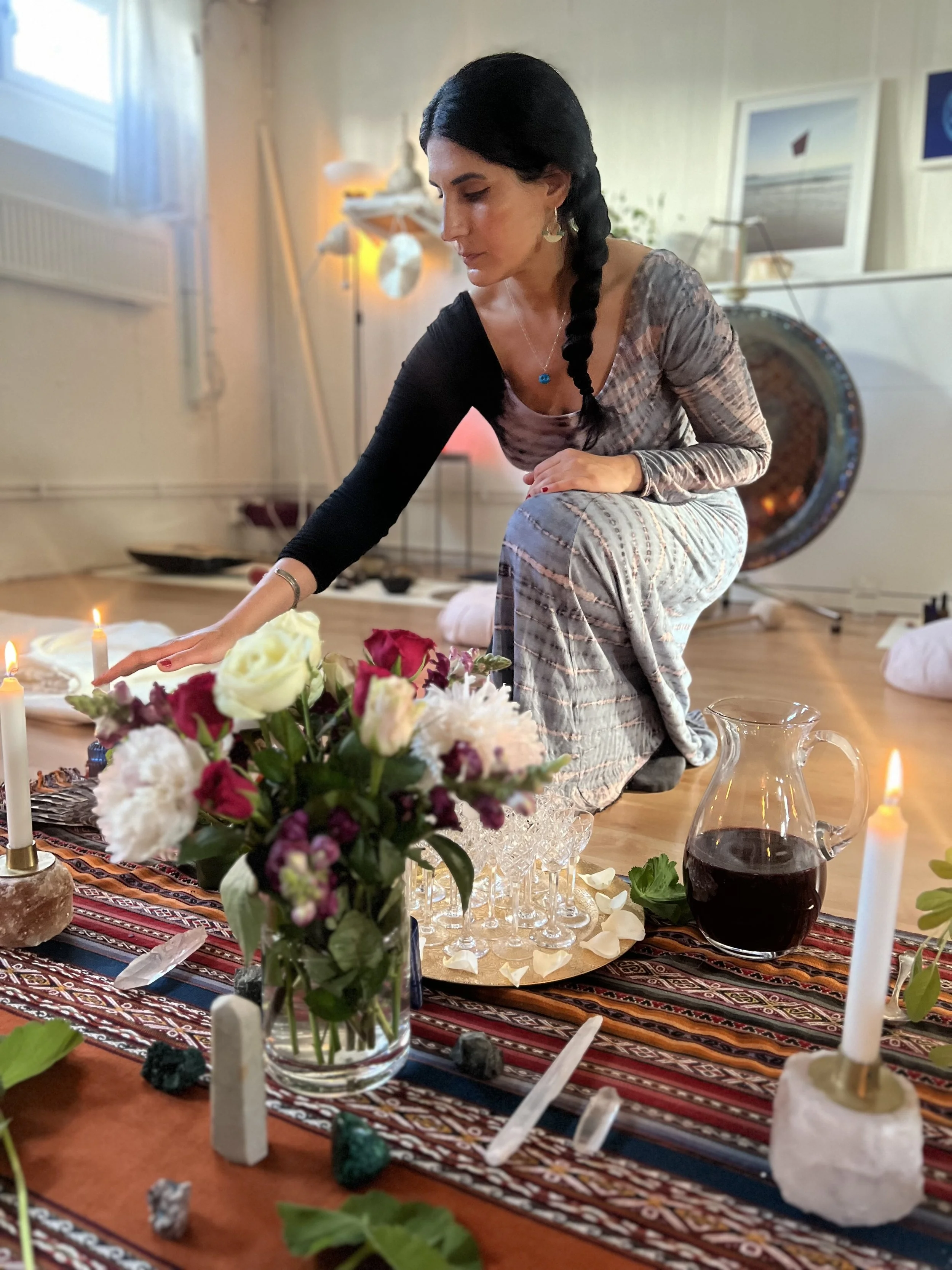 A woman with dark hair in a braid, dressed in a patterned dress, is kneeling on a cushion, reaching out to a flower arrangement on a table. The table has candles, a pitcher of red wine, and various ritual items, with a colorful cloth covering it. In 