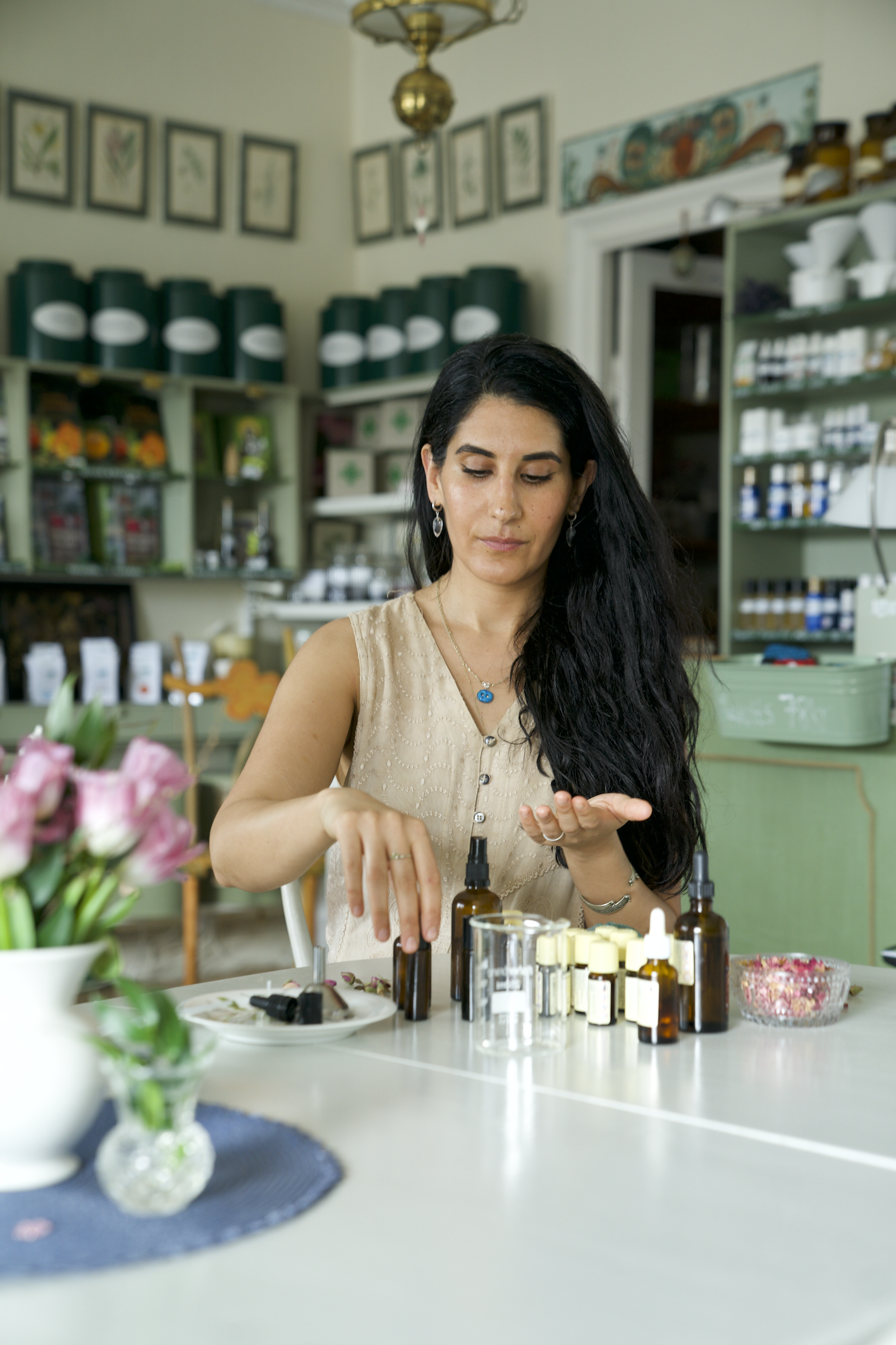Woman with long dark hair and earrings sitting at a table with bottles and a small dish, preparing a product, with a decorated room and shelves in the background.