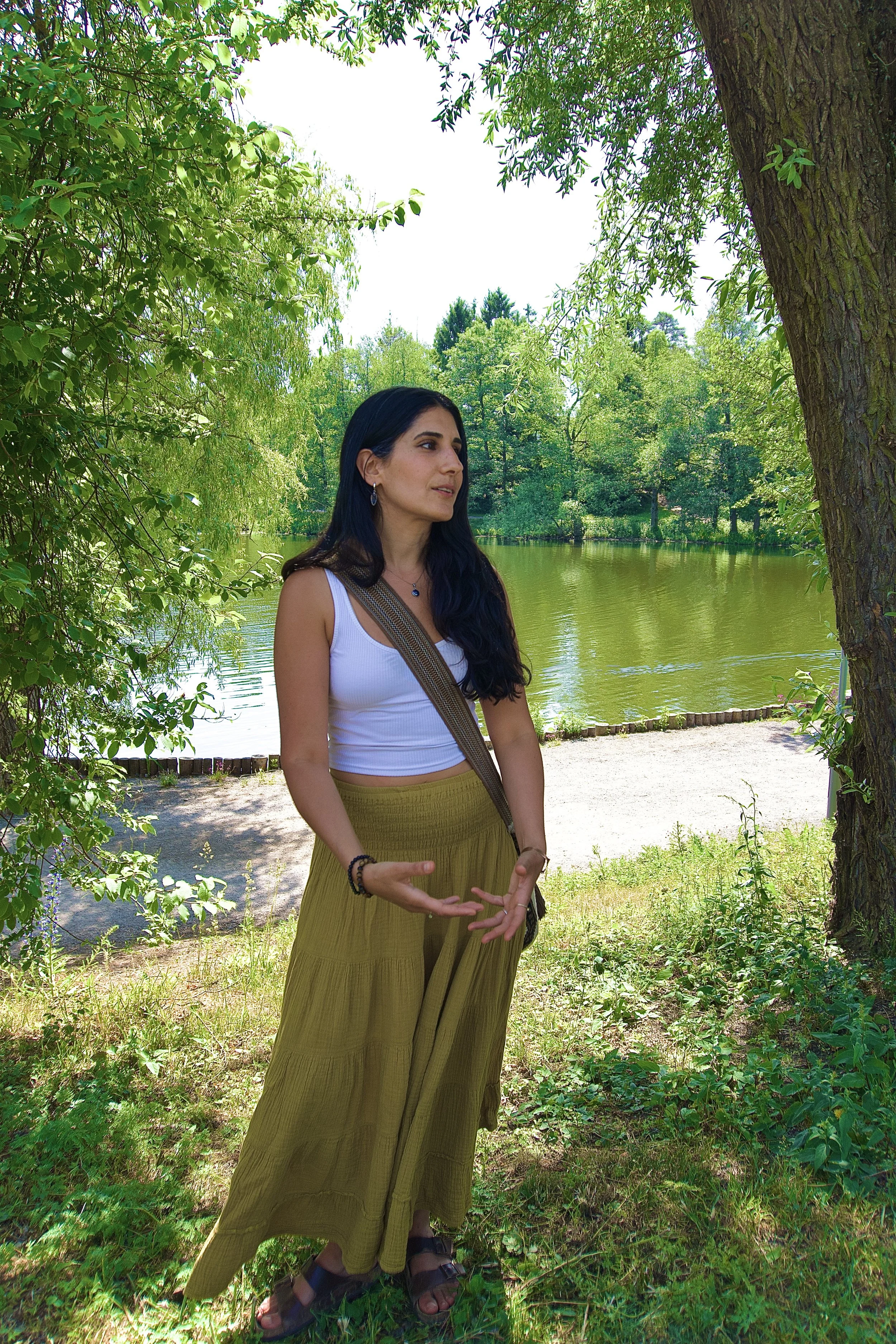 A woman with long dark hair standing under a tree near a pond, wearing a white tank top, mustard yellow wide-leg pants, and sandals, with a crossbody bag, surrounded by green foliage.