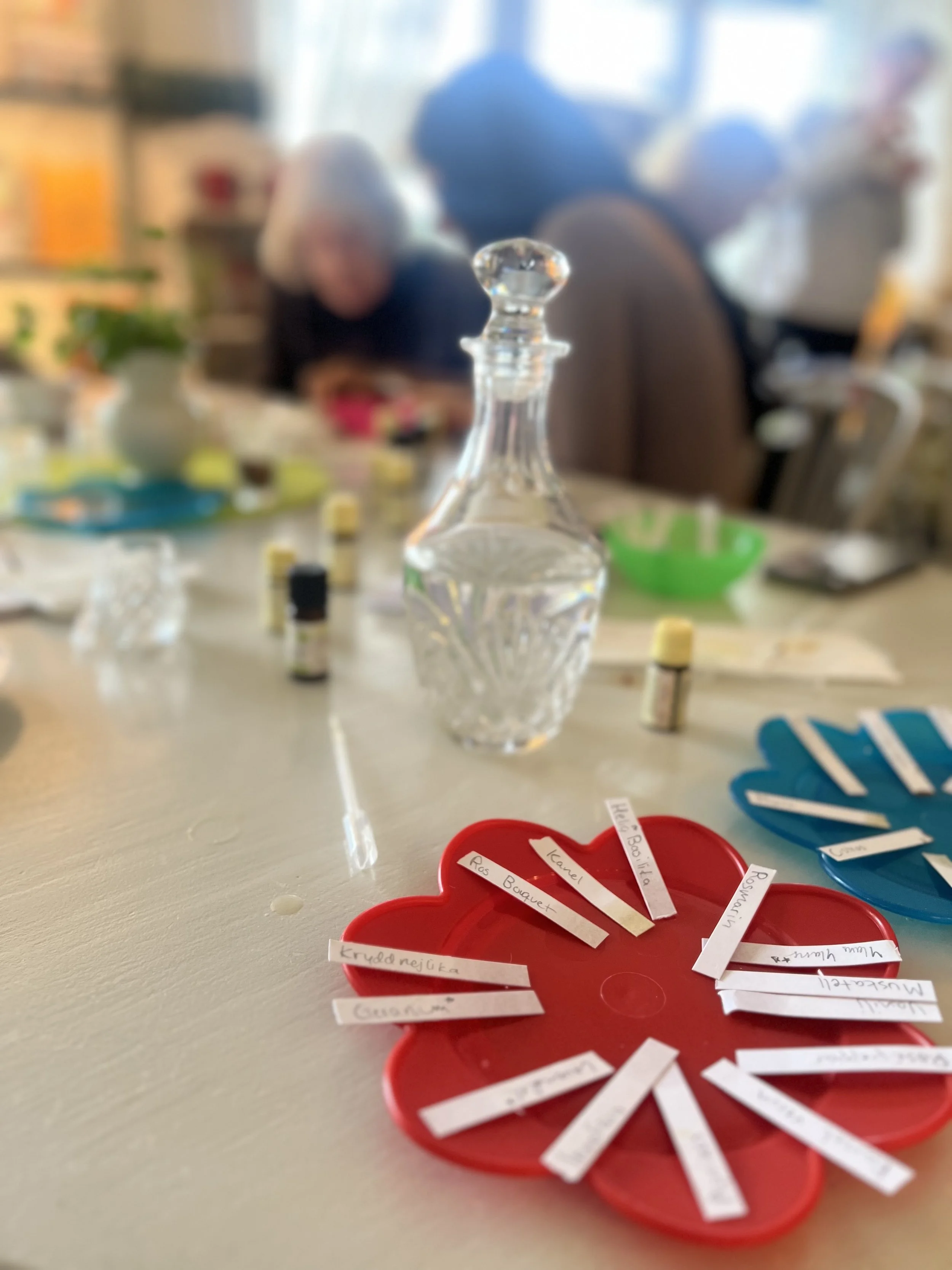 A tabletop with various items including a glass decanter, small bottles of essential oils, a green bowl, and a red flower-shaped container with small labeled paper strips, with people blurred in the background.