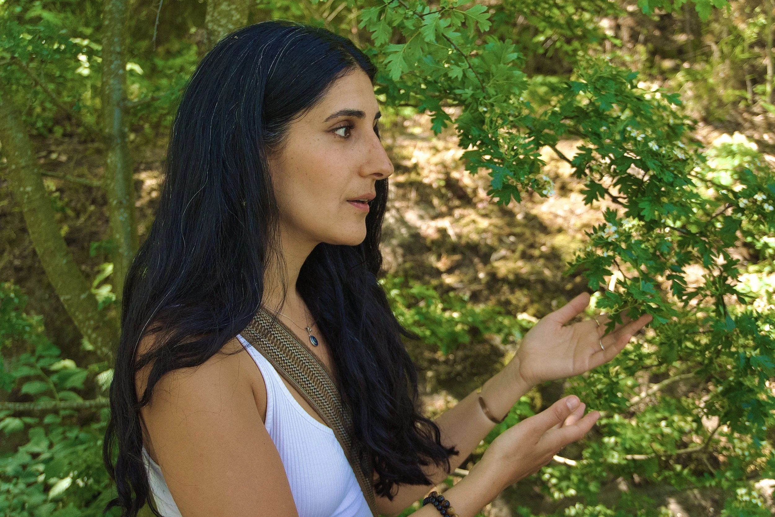 A woman with long dark hair and a white tank top standing in a lush green forest, touching the leaves of a tree.