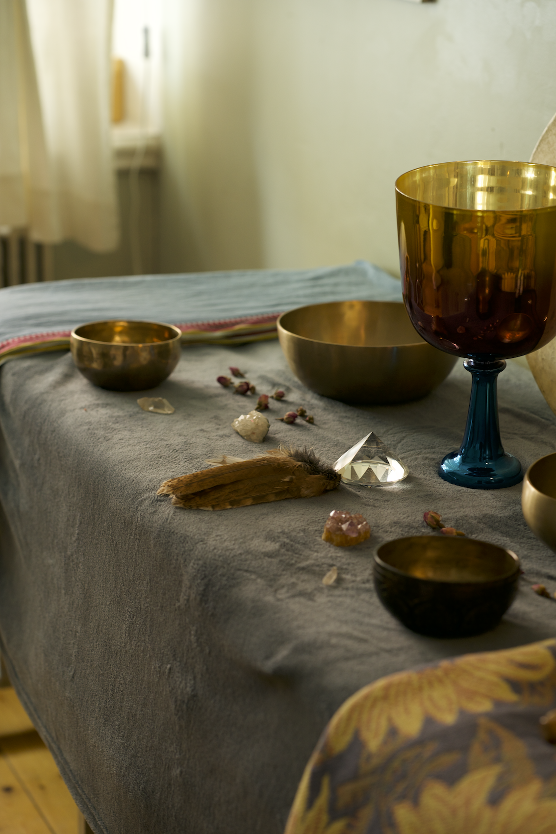 A table with a gray tablecloth decorated with bowls, crystals, feathers, and small stones or crystals, illuminated by natural light from a nearby window.