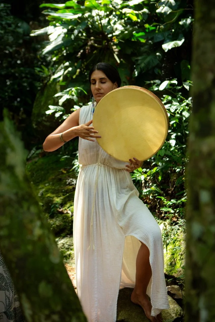 A woman in a white flowy dress is playing a large, round, yellow drum in a lush green forest.