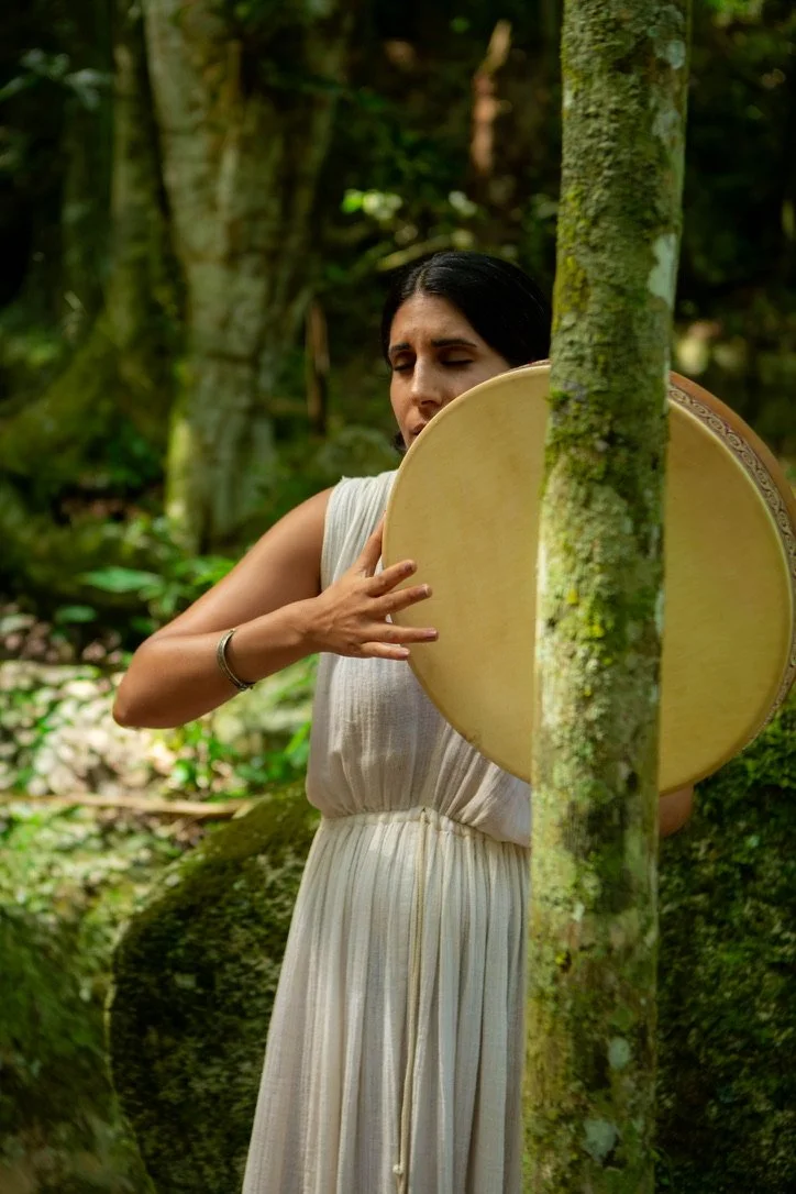 A woman in a white dress holding a large circular drum in a forest.