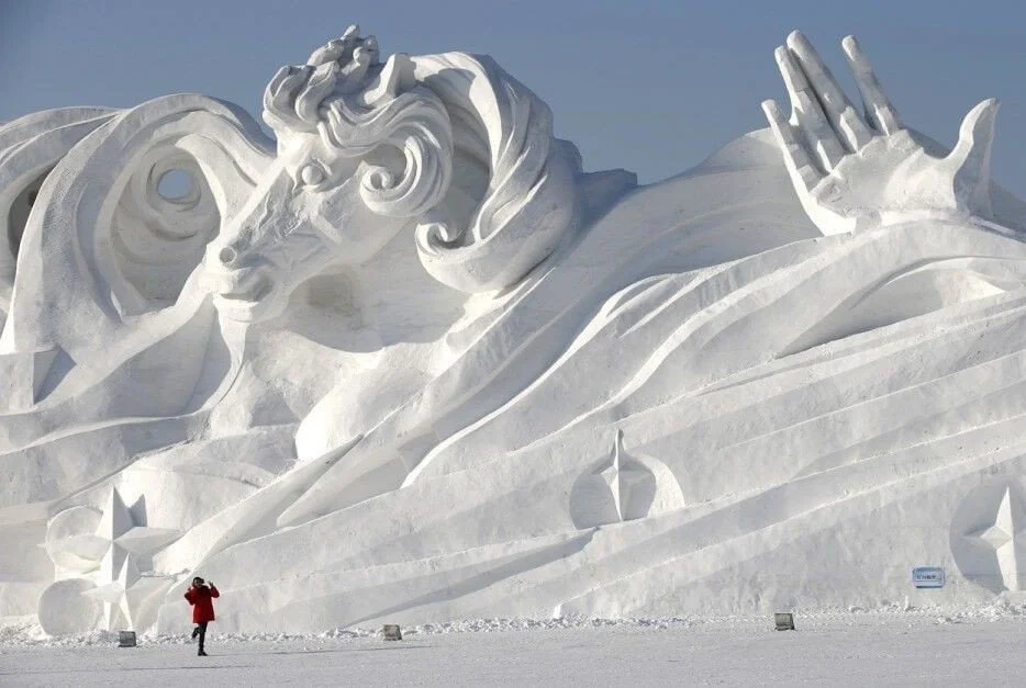 Large snow sculpture of a face with flowing hair and outstretched hand, set against a clear blue sky, with a person in red jacket taking a photo.
