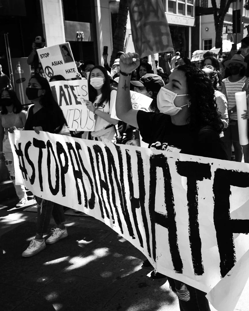 People participating in a protest, holding signs including one that says '#STOPASIANHATE' and another that says 'STOP ASIAN HATE'. Some are wearing masks.