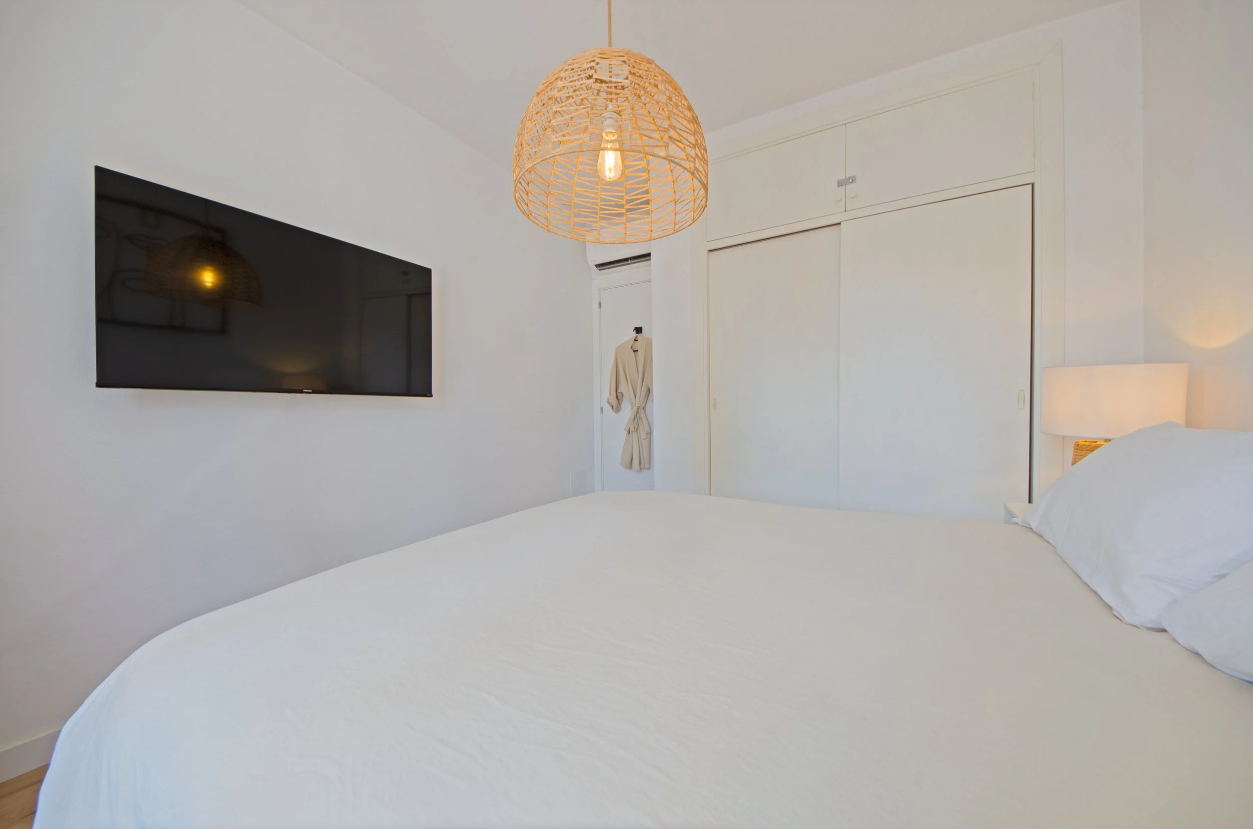 Cama con sábanas blancas, televisor en la pared, lámpara de mimbre y espejo en la habitación.