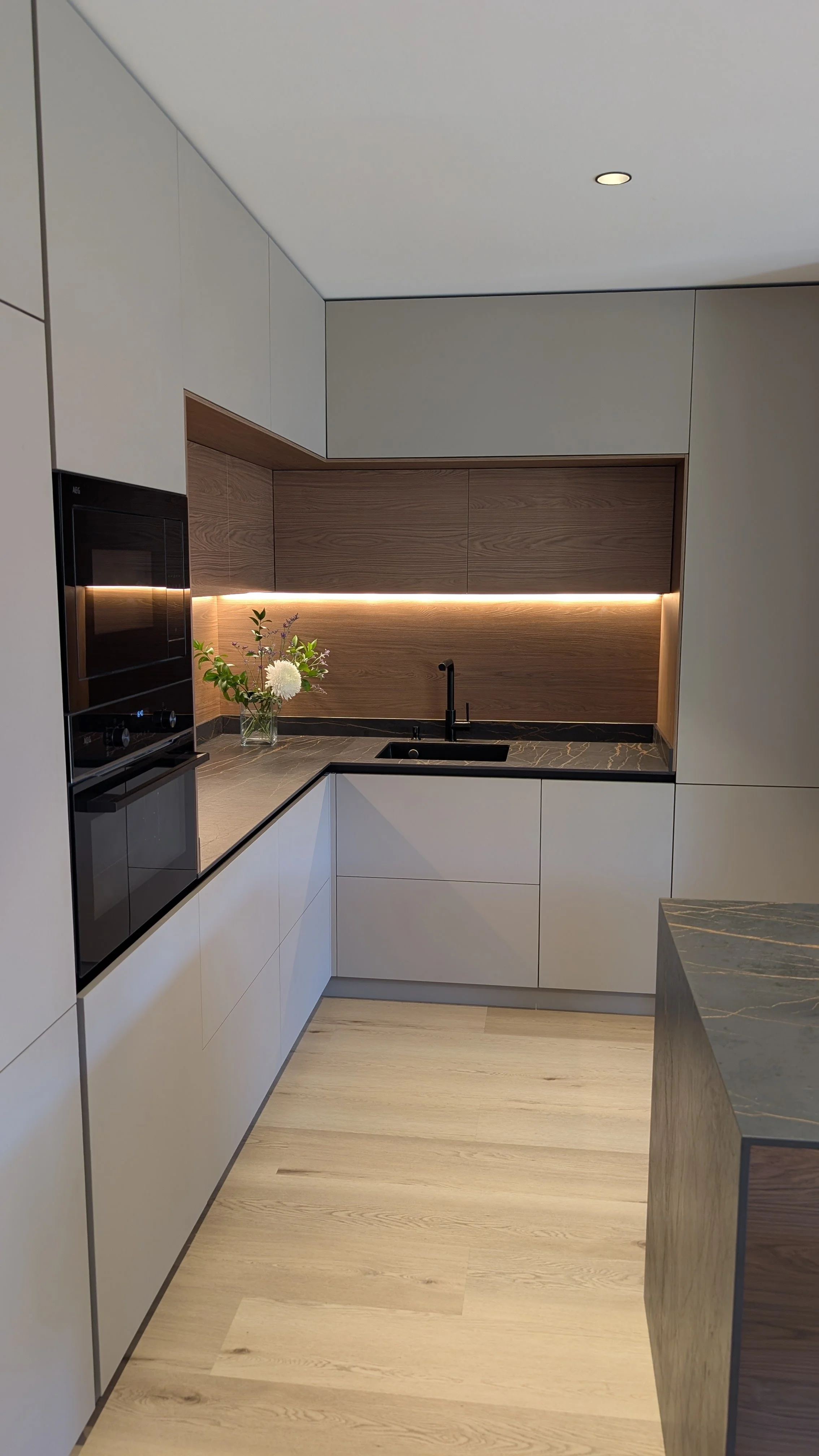 Cocina moderna con gabinetes blancos, encimera de mármol, fregadero negro, en la pared de fondo hay paneles de madera y una iluminación sutil debajo de ellos, decorada con un florero con flores blancas y verdes.