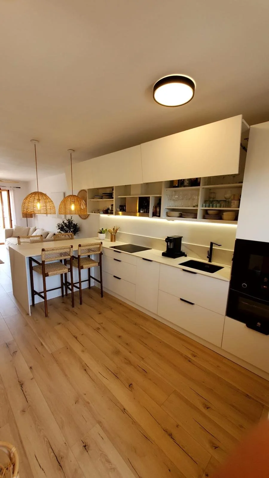 Cocina moderna con estantes abiertos, encimera de cocina blanca, iluminación LED, y una isla central con espacio para comer, decorada con plantas y utensilios de cocina diseñada y creada por seoane interiorismo en mallorca. 