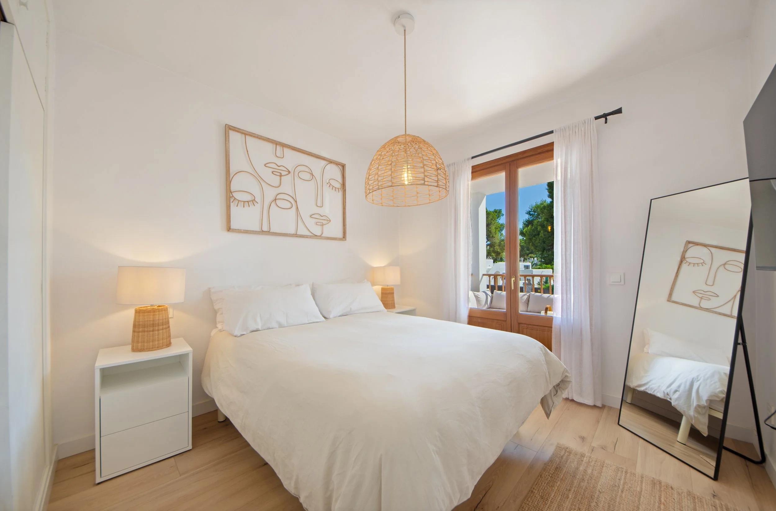 Dormitorio con cama blanca, cortinas blancas en una ventana de madera, lámpara de techo de mimbre, arte en la pared y almohadas blancas.
