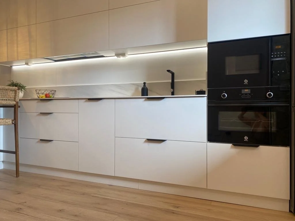 Cocina moderna con gabinetes blancos, encimera y pared de mosaico, horno empotrado negro, fregadero con grifo negro y detalles decorativos minimalistas diseñada por seoane interiorismo en mallorca. 
