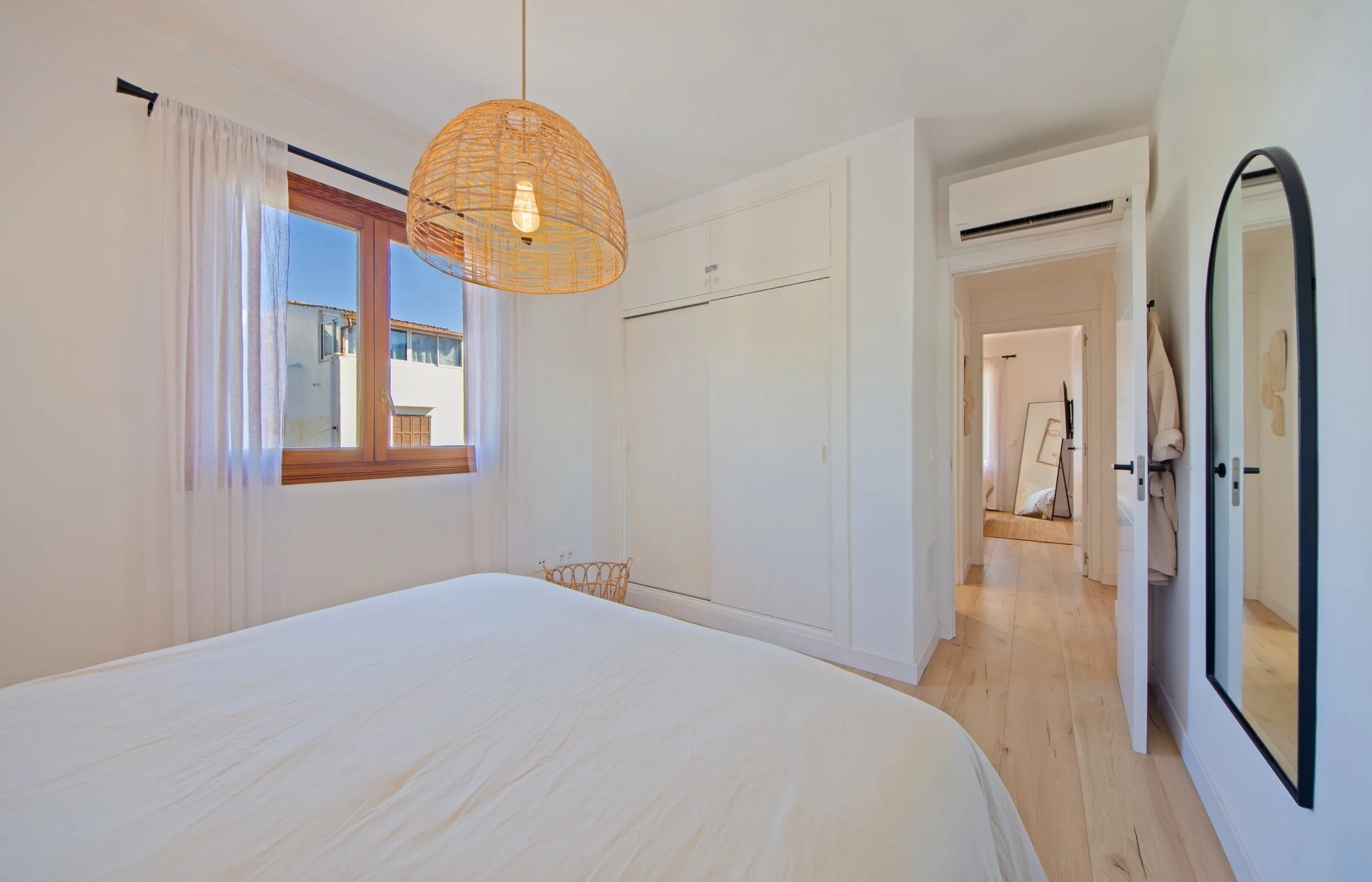 Habitación con paredes blancas, ventana de madera con cortinas blancas, lámpara de mimbre, puertas blancas, aire acondicionado y espejo de cuerpo completo.